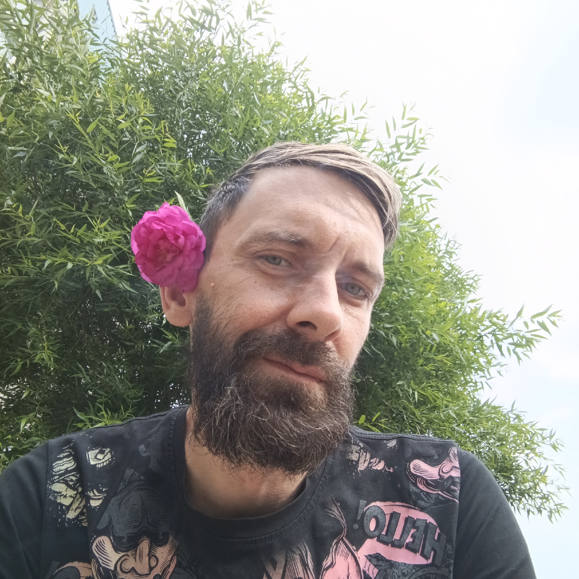 A man with light brown hair, a beard, and a pink flower tucked behind his left ear is shown in a close-up, upper body selfie. He is wearing a black t-shirt with a graphic print. He is outdoors, with a blurry green foliage and a bright sky in the background. The man is looking towards the camera with a slight smile. The location is stated to be Марковский, Russia.