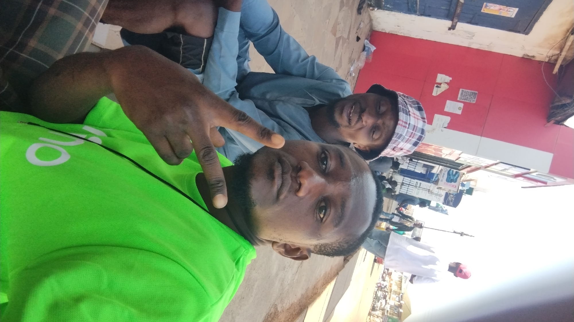 Two dark-skinned men are posing for a selfie-style photograph. The man in the foreground is wearing a bright green shirt and making a hand gesture with his fingers. The other man is wearing a light blue shirt and a checkered hat. They appear to be outdoors in Jos, Nigeria, based on background details. There are buildings with advertisements and other people in the background, suggesting a commercial area. The image is slightly rotated to the side.