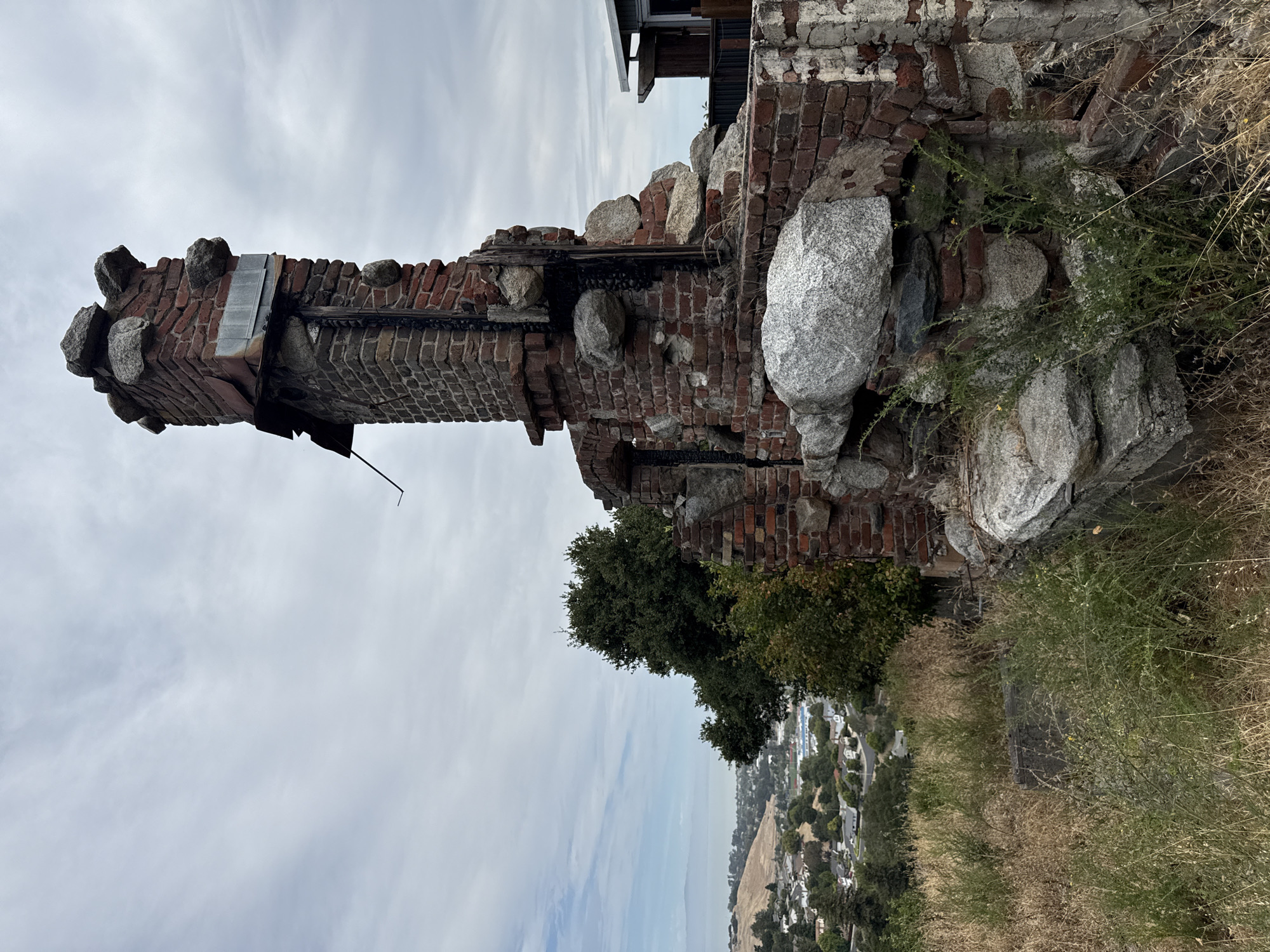 A weathered brick and stone chimney structure stands on a hilltop overlooking a suburban landscape in El Sobrante, California, USA. The chimney is partially collapsed and appears to be part of a former building. The brickwork is red and aged, with some sections missing. Large, rounded stones are incorporated into the structure, some protruding outwards. The top of the chimney is capped with similarly sized stones. Visible charring and soot are present on the brickwork, suggesting the structure may have been involved in a fire. Surrounding the chimney is dry, overgrown vegetation, including tall grasses and bushes. In the background, houses and trees of a residential area are visible, leading down towards a body of water in the distance. The sky is overcast with scattered clouds. The overall scene conveys a sense of decay and abandonment.