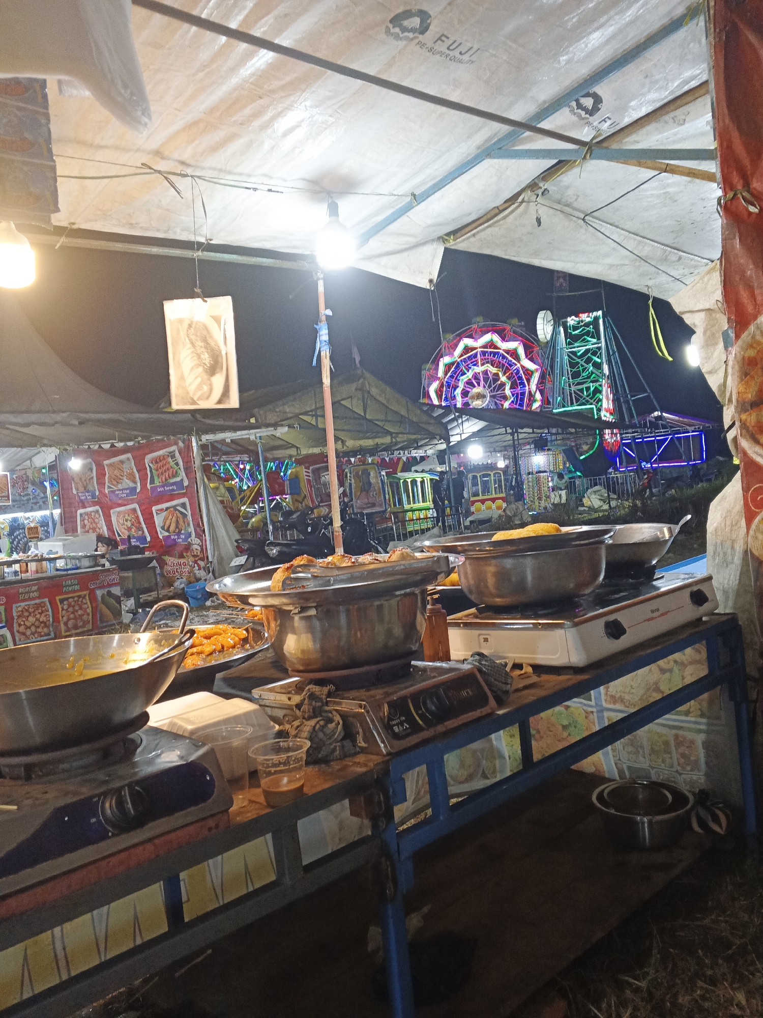The image depicts a night scene at what appears to be an outdoor food stall and carnival at a market in Sukabumi, Indonesia. In the foreground, a metal table holds cooking equipment, including a portable gas stove with two burners, several large metal pots and pans, and a tray of fried food. One pot contains a yellow liquid, likely a sauce or curry. To the right of the stove are two plastic cups, one containing a beverage, and a crumpled cloth.
Behind the food stall, a vibrant, illuminated Ferris wheel and other carnival rides are visible in the background, set against a dark night sky. Tents and tarpaulins, supported by metal frames and bamboo poles, provide shelter over the stalls. Various advertisements are displayed on posters and banners, some featuring food items. A motorcycle is parked behind one of the stalls. The ground is covered with dry grass or straw. The lighting suggests the presence of overhead bulbs illuminating the area.