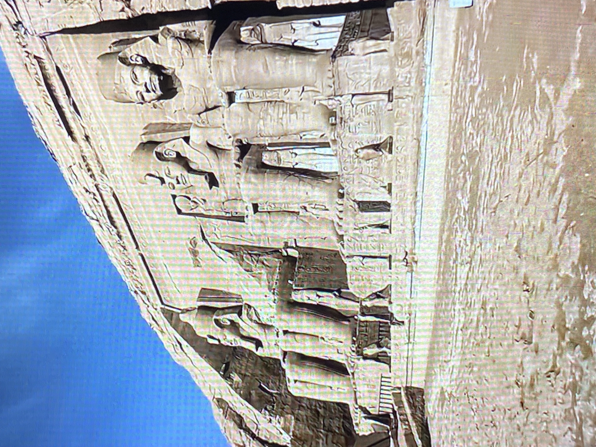 The image shows the façade of the Great Temple of Ramses II at Abu Simbel, Egypt. The temple is carved into a sandstone cliff face, with four colossal statues of Ramses II seated at the entrance. Hieroglyphic inscriptions cover the walls around the statues. The left side of the image shows the deep blue of the sky. The right shows the rock cliff. A metal ladder is visible in the bottom right. The image is shown rotated counter-clockwise 90 degrees. This structure is not located in Tinaquillo, Venezuela.