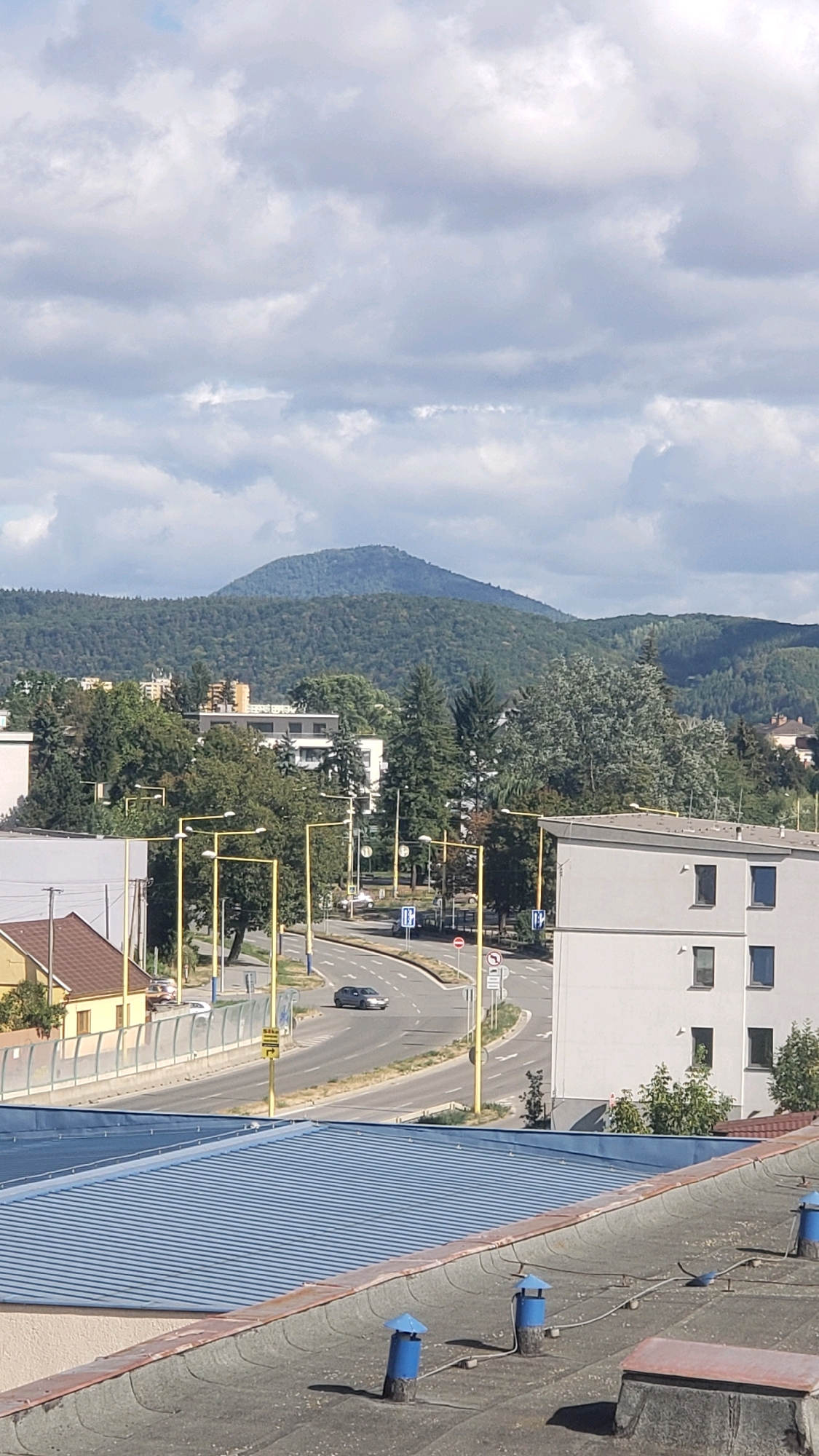 The image shows a panoramic view of a city street and buildings with a mountain range in the background under a cloudy sky. The scene appears to be in the city of Prešov, Slovakia, with the iconic Zemplín Castle ruins visible on a distant hill. In the foreground, a rooftop with ventilation pipes and a blue corrugated metal roof is visible. Beyond that, a street curves through the urban landscape, with cars, traffic lights, and street lamps lining the road. Several buildings of various sizes and architectural styles are present, including residential buildings and what appears to be a commercial property. The mountain range, covered in dense green trees, dominates the horizon, with a distinct peak in the center. The sky is filled with a mix of white and gray clouds, suggesting a partly cloudy day. The overall impression is that of a typical urban environment nestled within a natural landscape.