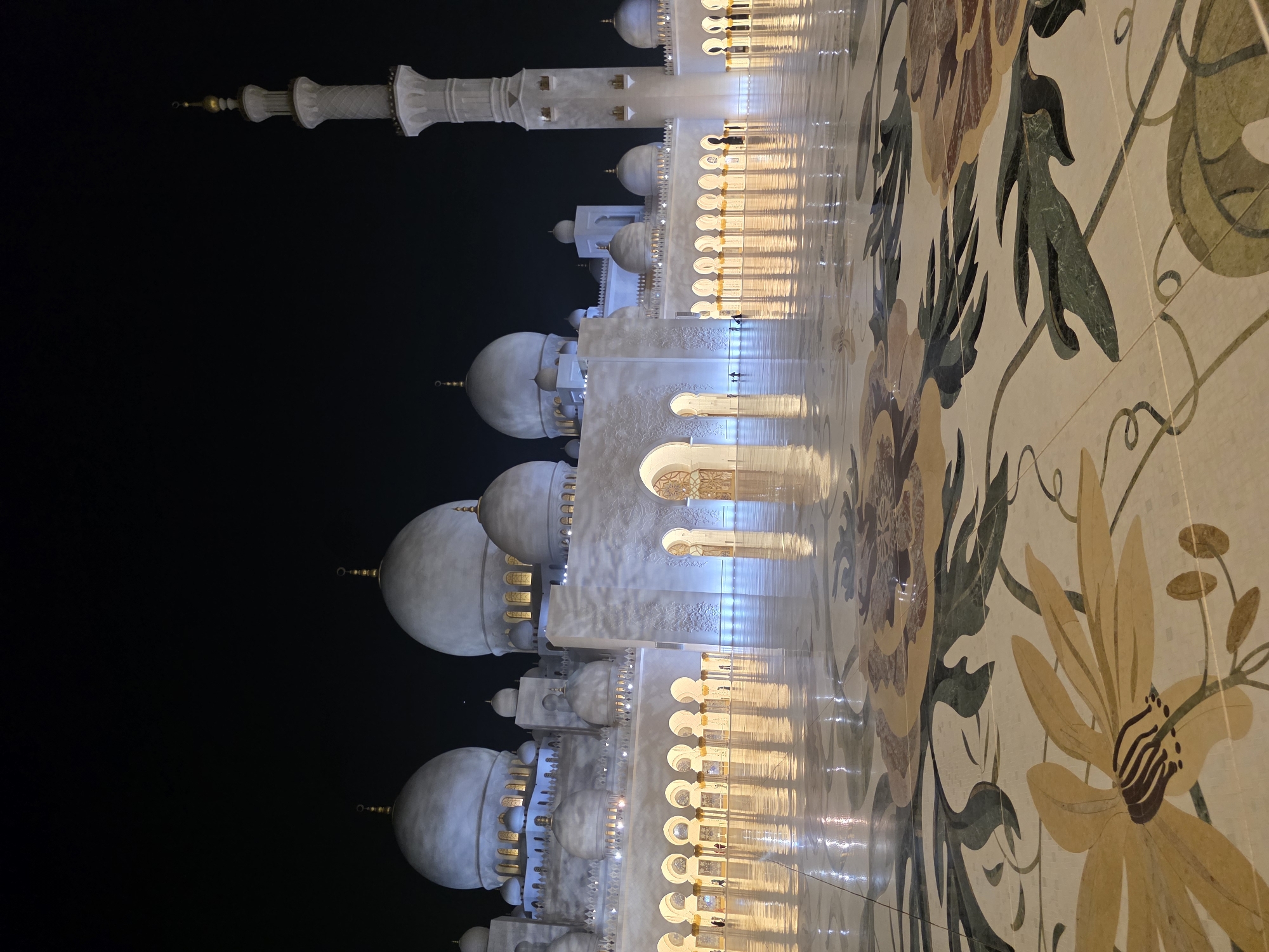 The image captures a nighttime view of the Sheikh Zayed Grand Mosque in Abu Dhabi, United Arab Emirates. The mosque's white marble domes and minarets are illuminated against the dark sky. The foreground features a highly polished floor with an intricate floral mosaic pattern, reflecting the lights of the mosque. Several arched walkways with columns lead into the structure. A few silhouetted figures can be seen walking in the distance, along the arcades. The overall scene conveys a sense of grandeur and tranquility.