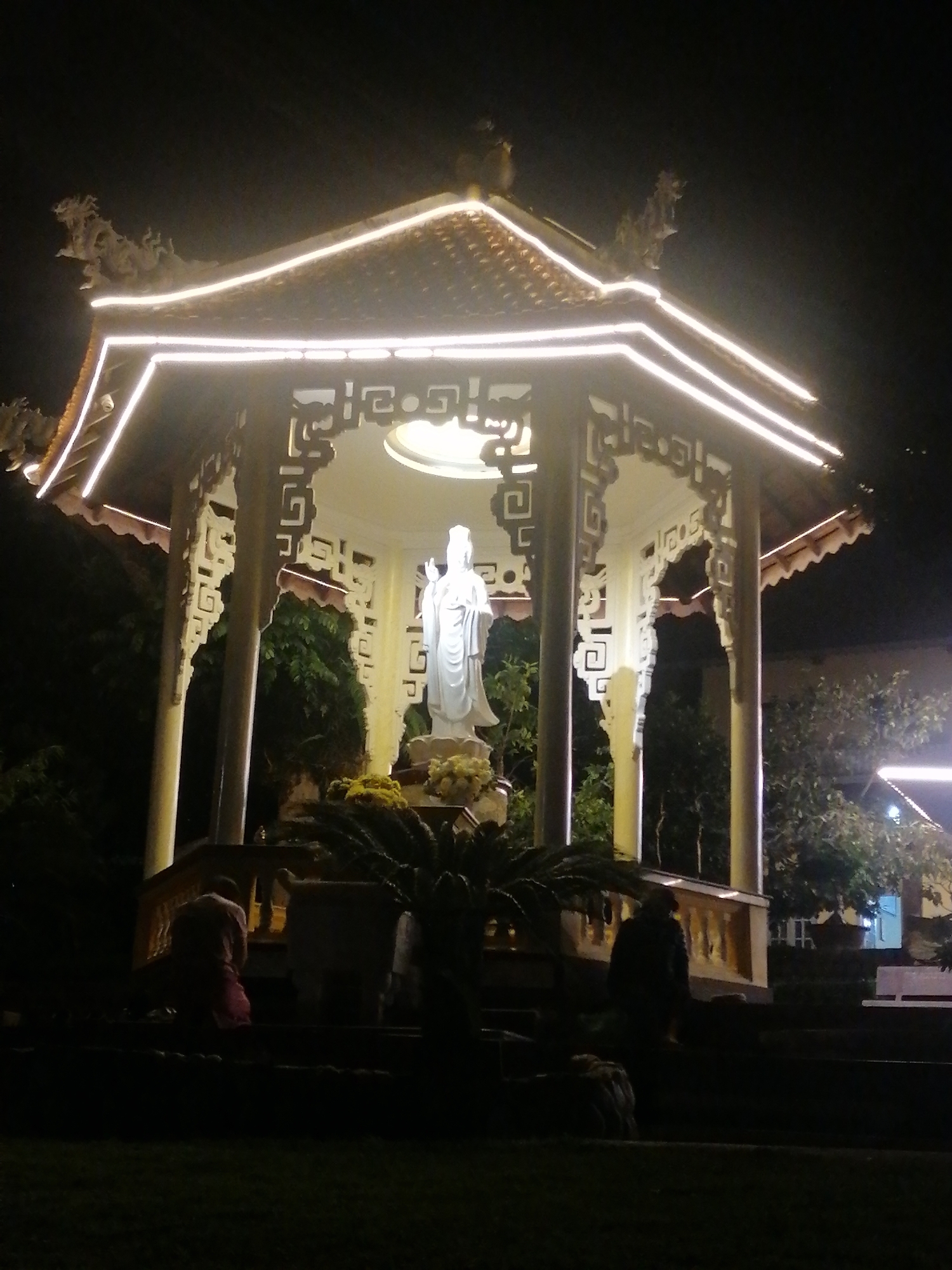 The image depicts a brightly lit pagoda at night, possibly located in Da Lat, Vietnam. A statue of a standing female figure, possibly a deity, is centrally positioned within the pagoda's illuminated interior. The pagoda has a tiered roof adorned with decorative carvings and is outlined with white LED strip lights. Several figures are visible in silhouette on the steps leading up to the pagoda, some appearing to be kneeling or walking. The surrounding environment is dark, with trees and foliage visible in the background, suggesting an outdoor setting.