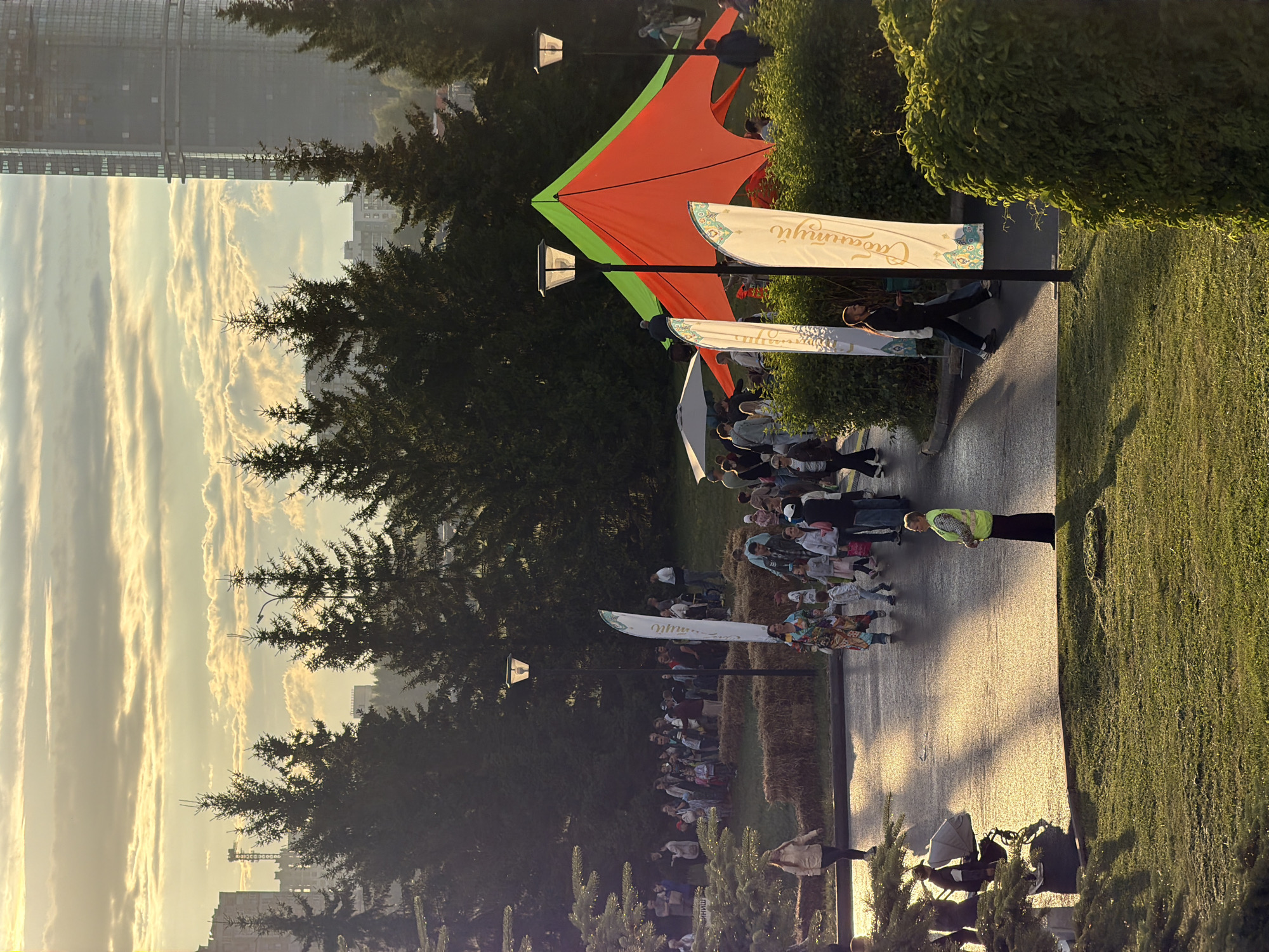 The image depicts an outdoor event at dusk, likely in a park, with a crowd of people gathered. In the background, tall evergreen trees frame a city skyline featuring buildings and a prominent glass skyscraper under construction. The sky is filled with soft, illuminated clouds, and the setting sun casts long shadows and a warm glow on the wet asphalt pathway.
In the foreground, a lush green lawn slopes gently upwards, with a person in a high-visibility vest standing on it. The pathway is populated by many individuals, including families with children and adults walking around. A bright orange and green tent is set up, and several tall, white flags with text and ornate designs are planted nearby. Hay bales are stacked in a few areas, possibly as seating or decoration. A woman in a colorful patterned dress is walking on the pathway, and a child is walking beside her. Another woman in a light-colored jacket is walking on the pathway away from the viewer. Some trees with feathery needles are visible in the lower left corner, casting shadows on the ground. Two lampposts with modern, solar-powered lights are present. The scene suggests a lively community gathering.