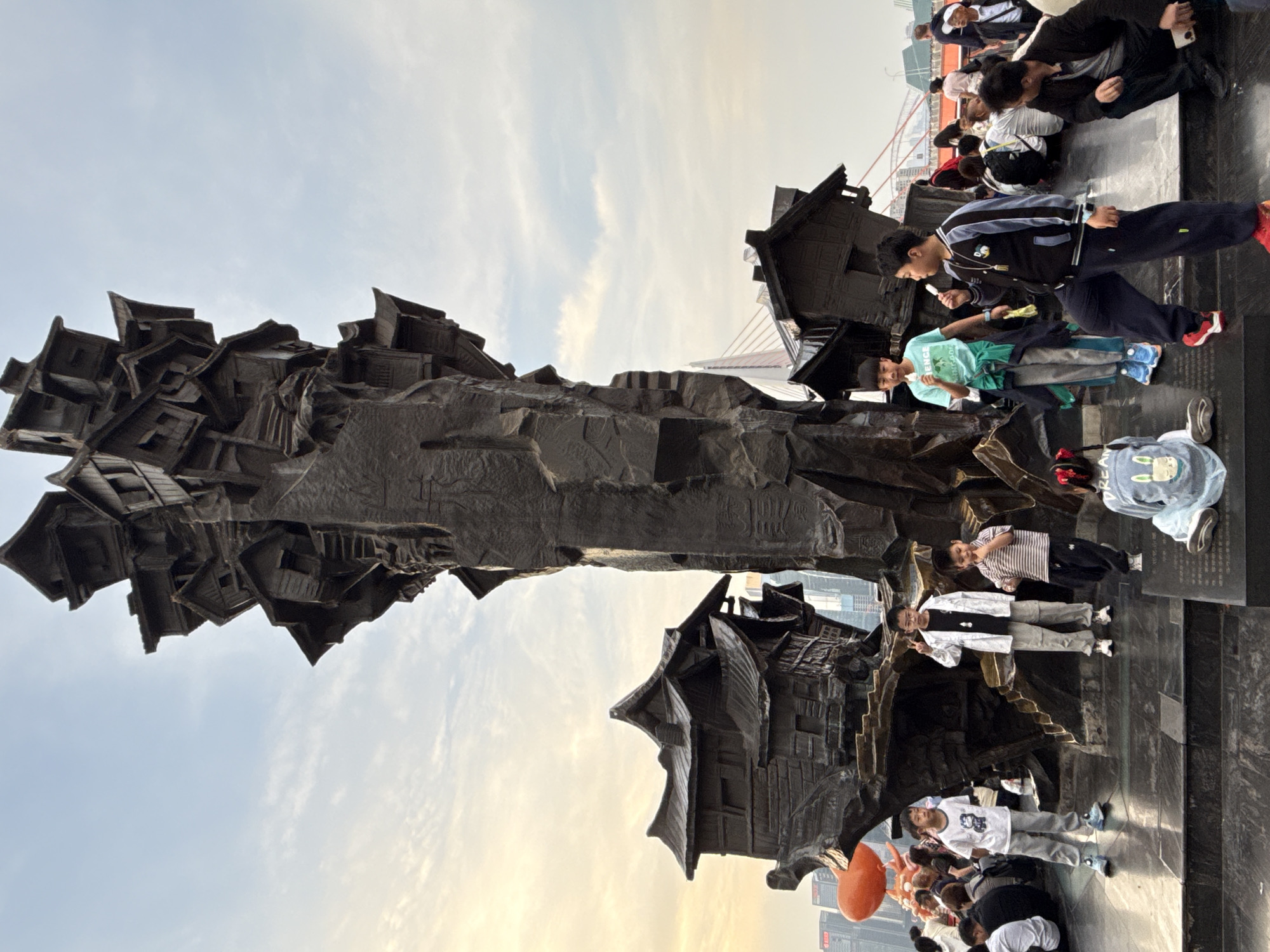 The image shows a large, dark-colored sculpture in an outdoor setting. The sculpture appears to be made of stone or metal and is composed of multiple house-like structures stacked together.
Several people are interacting with the sculpture. Some are standing or sitting nearby, while others appear to be climbing on or posing with the sculpture for photos.
The background includes a cityscape with buildings and a bridge visible in the distance. The sky is partly cloudy. The location is Yuzhong District, China.