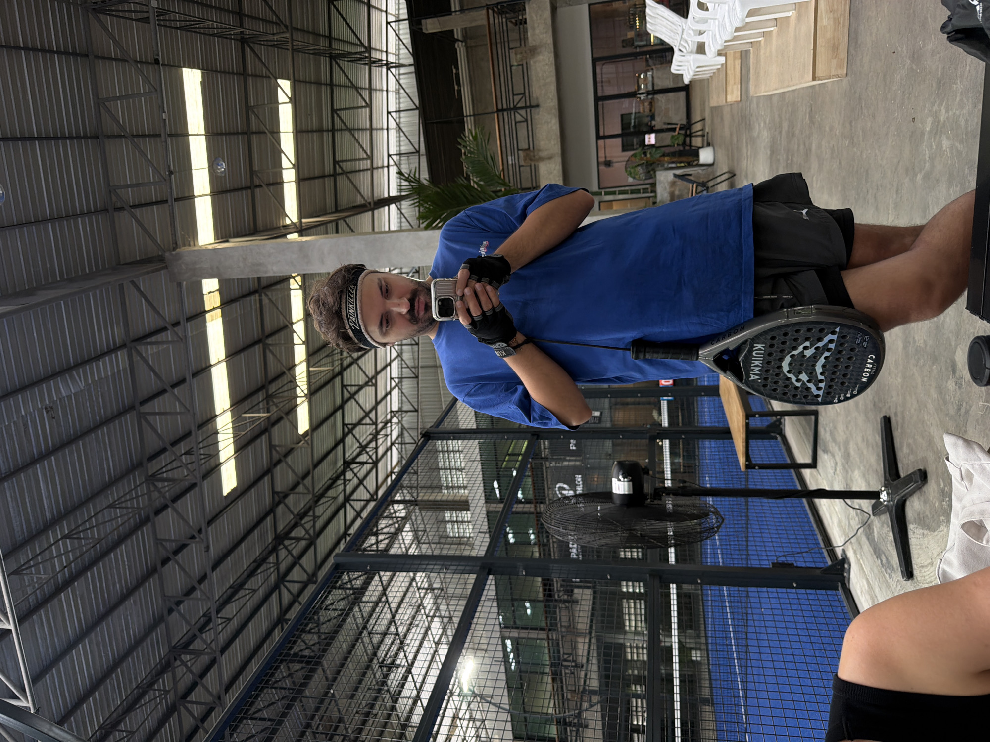 The image depicts a person holding a phone and a paddle. The person is wearing a blue t-shirt, black shorts, a black headband, and black gloves. They are standing on a concrete floor, with a large fan and a blue court visible in the background. The setting appears to be an indoor sports facility with a high, corrugated metal ceiling and exposed metal beams. The lighting suggests it could be daytime. The person is looking at their phone, likely taking a selfie or recording a video. The paddle has "KUIMIA" and "CARBON" printed on it. The scene is well-lit, with sunlight streaming through gaps in the roof. The overall atmosphere seems casual and sporty. The headband has text that is partially visible and appears to read "PAINKILLER".