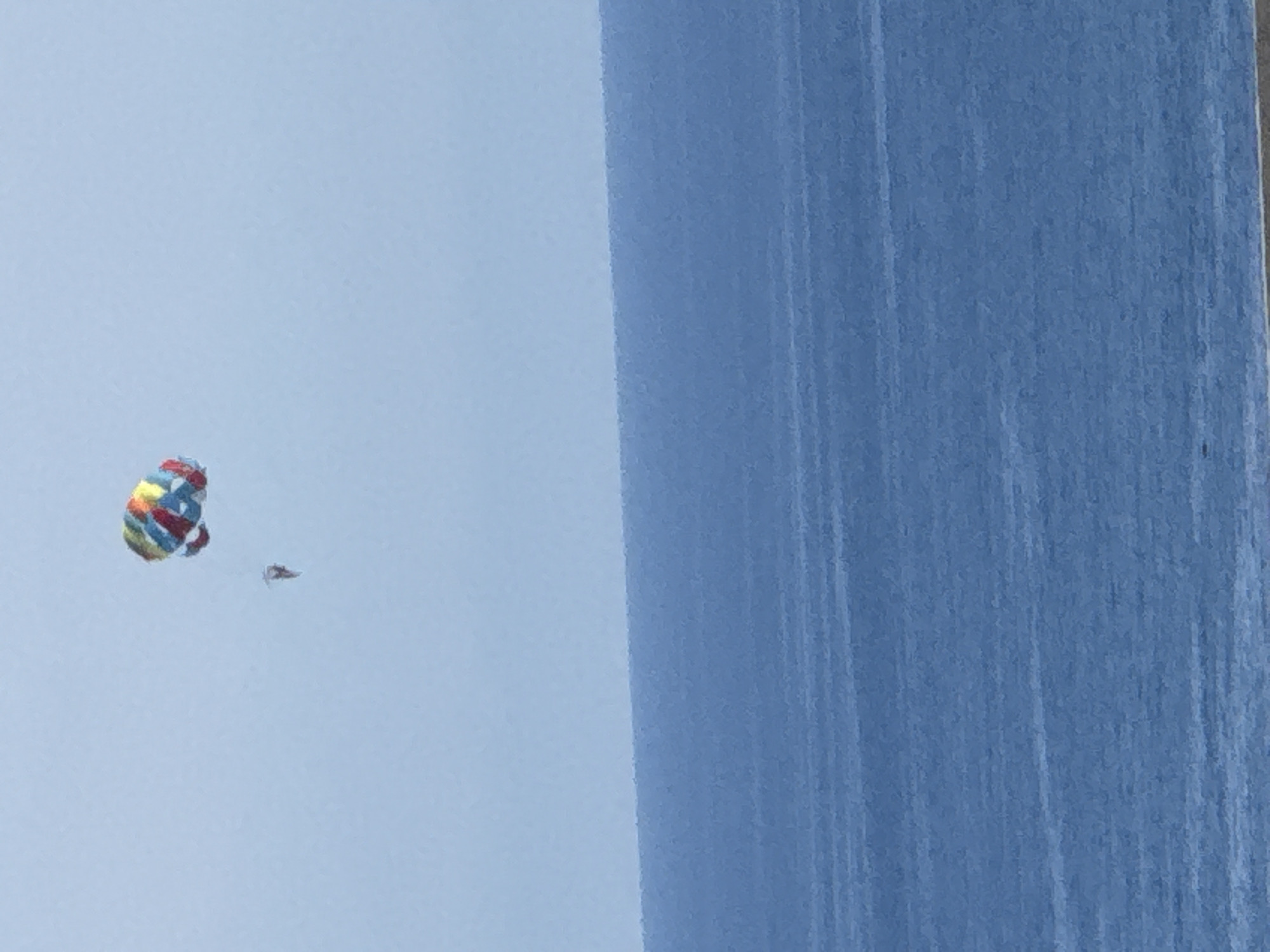 The image captures a serene scene of a body of water, likely the Adriatic Sea, with a clear, light blue sky above. In the sky, a colorful parasail, adorned with red, yellow, green, and blue hues, is visible. Attached to the parasail is a person, though their features are indistinct due to the distance. The water is a deep, consistent blue, with subtle horizontal lines suggesting gentle waves. The horizon line where the water meets the sky is sharp. The image quality is somewhat grainy. The location is identified as Boreti, Montenegro, a coastal area known for its beaches and clear waters, making parasailing a popular activity.