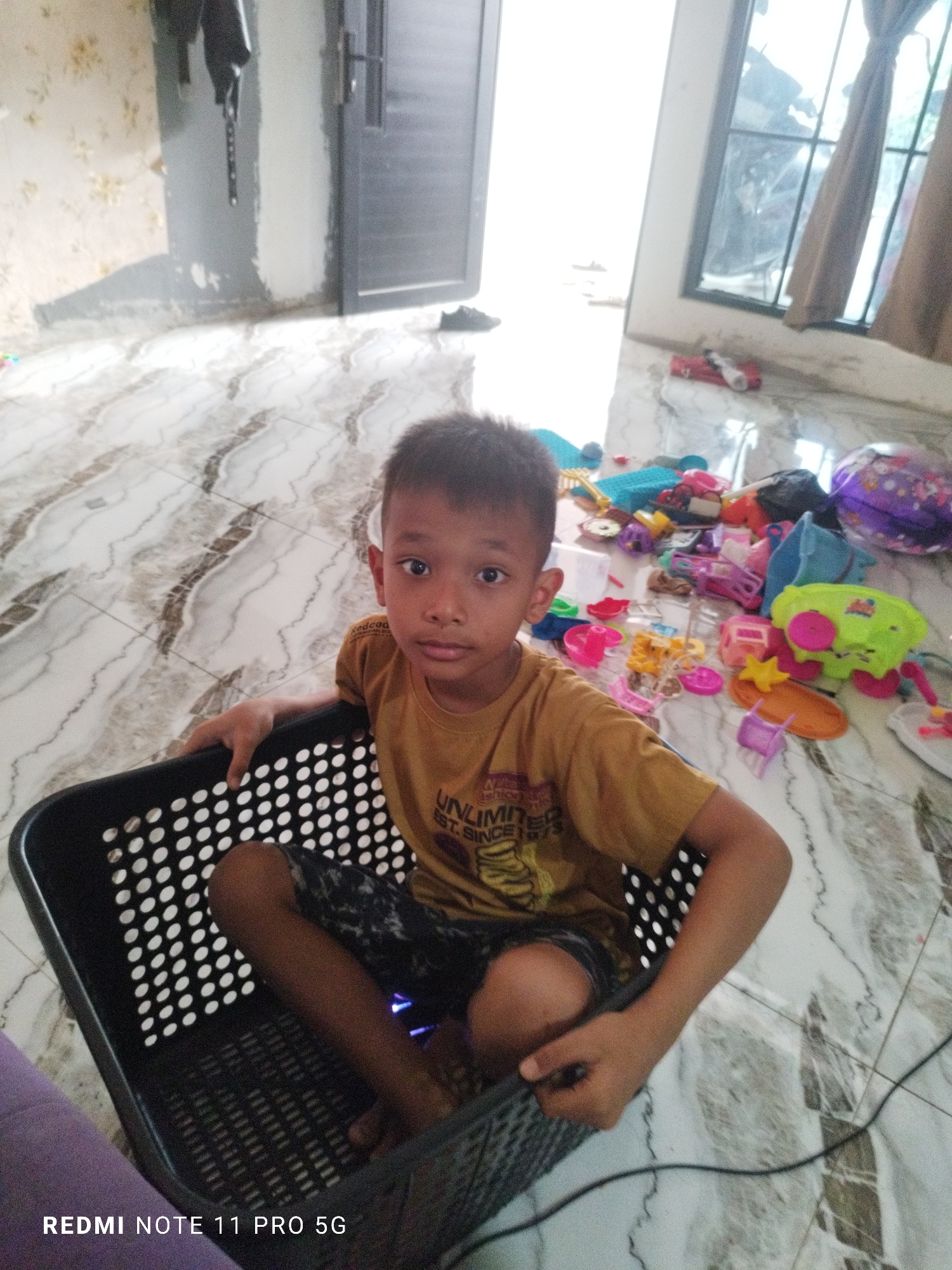 A young Indonesian boy, likely under ten years old, sits in a black plastic laundry basket in a room with marble-patterned flooring. He is wearing a yellow t-shirt with a graphic that reads "UNLIMITED SINCE 1973" and patterned shorts. His left arm rests on the edge of the basket, and his right hand is in his lap. The room appears to be cluttered with various toys scattered on the floor, including brightly colored plastic toys, a purple helmet, and other items. A doorway is visible in the background to the left, and a window covered by curtains is to the right. The overall lighting suggests it is daytime, possibly with sunlight streaming in. The setting is consistent with a residential dwelling in Kaduhejo, Indonesia.