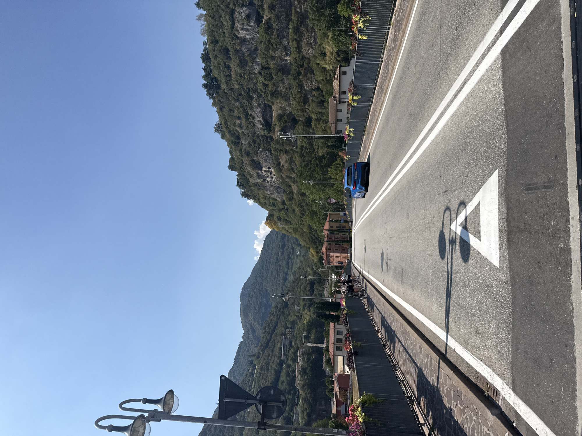 The image captures a view from a bridge in the city of Trento, Italy. The bridge is paved with asphalt and marked with white lines, indicating lanes for vehicles. A blue car is driving away from the camera on the right lane. On the left side of the bridge, there is a sidewalk with a metal fence and flower boxes. Several people are walking their bicycles on the sidewalk, facing away from the camera. Beyond the bridge, the landscape is dominated by lush green mountains with some scattered buildings on their slopes. The sky is clear and blue. The scene appears to be a sunny day, likely in the afternoon given the shadows.