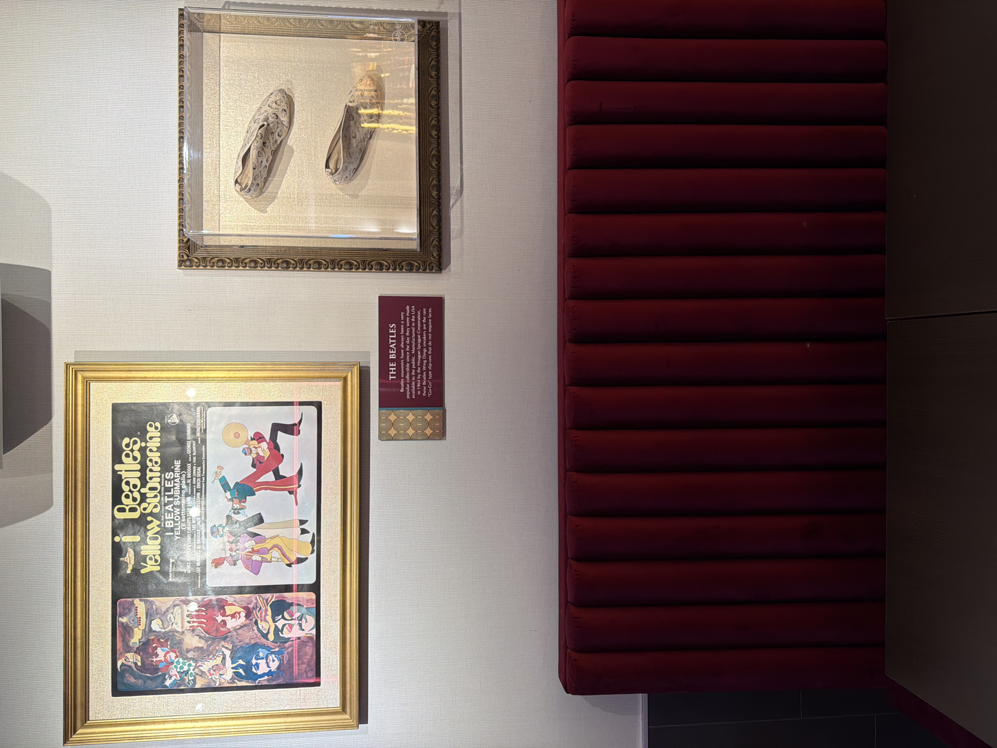 This indoor image presents a display of Beatles memorabilia against a light-colored wall, indicative of a museum or exhibition setting.
On the upper left, a pair of light-colored, patterned slip-on shoes are encased in a clear box within a decorative gold frame. Below this, a larger gold frame showcases a colorful, psychedelic poster for "The Beatles Yellow Submarine" animated film. The poster prominently features the title "iBeatles Yellow Submarine" and stylized illustrations of the band members and characters. Credits for individuals like "GEORGE DUNNING," "JOHN LENNON," "PAUL McCARTNEY," and "HEINZ EDELMANN" are visible.
Between the two framed items, a small, dark red informational plaque titled "THE BEATLES" offers details about the displayed footwear. The text explains, "Beatles souvenirs have always been a very popular collectible since the day they were made available to the public. Manufactured in the USA in 1964 by the Hoague-Sprague Corporation, these Beatles Wing Dings sneakers are the rare 'Go-Go' type slip-ons that do not require laces." This confirms the shoes are original Beatles merchandise.
To the right of these displays, a large section of dark red, horizontally ribbed upholstered material extends vertically, possibly serving as a decorative wall panel or part of a seating area. A narrow, dark vertical panel borders the right edge of the frame. The scene is illuminated by artificial indoor lighting, enhancing the details of the exhibited artifacts. The overall ambiance suggests a dedicated space for pop culture and music history appreciation, potentially located in Milan, Italy.