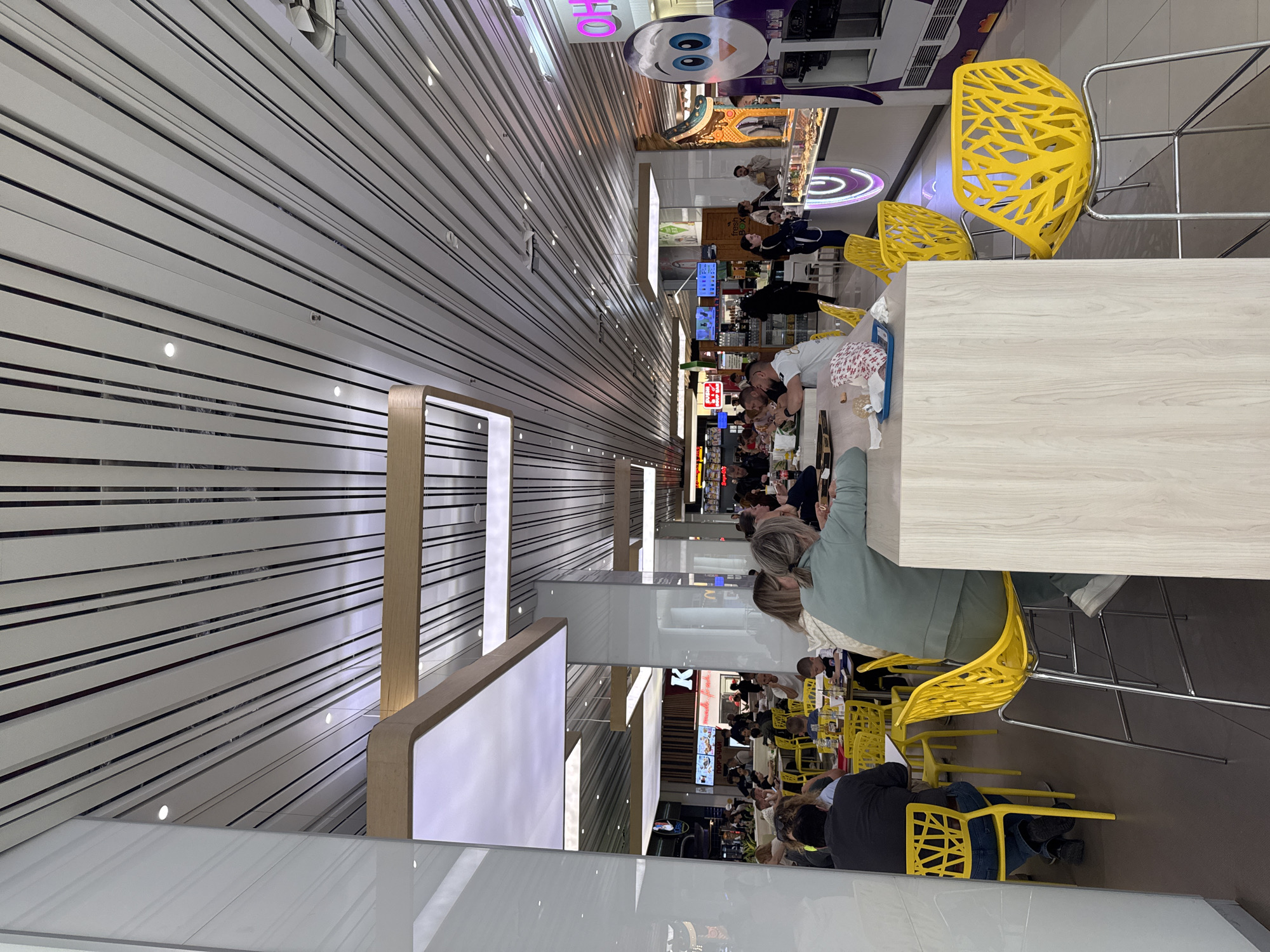 The image depicts the interior of a food court area, presumably in a mall or airport, given the scale and design. The location is likely Plovdiv, Bulgaria, though there are no direct visual cues to confirm this. The scene is characterized by rows of tables and chairs. People are sitting at the tables, presumably eating or resting. A few are clustered together, possibly in conversation. The seating consists of bright yellow, geometrically-designed chairs, some are high stools with metal frames. The tables are primarily a light wood-grain finish. There are illuminated white panels hanging from the ceiling. The ceiling is constructed from white and dark narrow, horizontal panels, interspersed with small, recessed lights. In the background, there are various storefronts and food vendors.