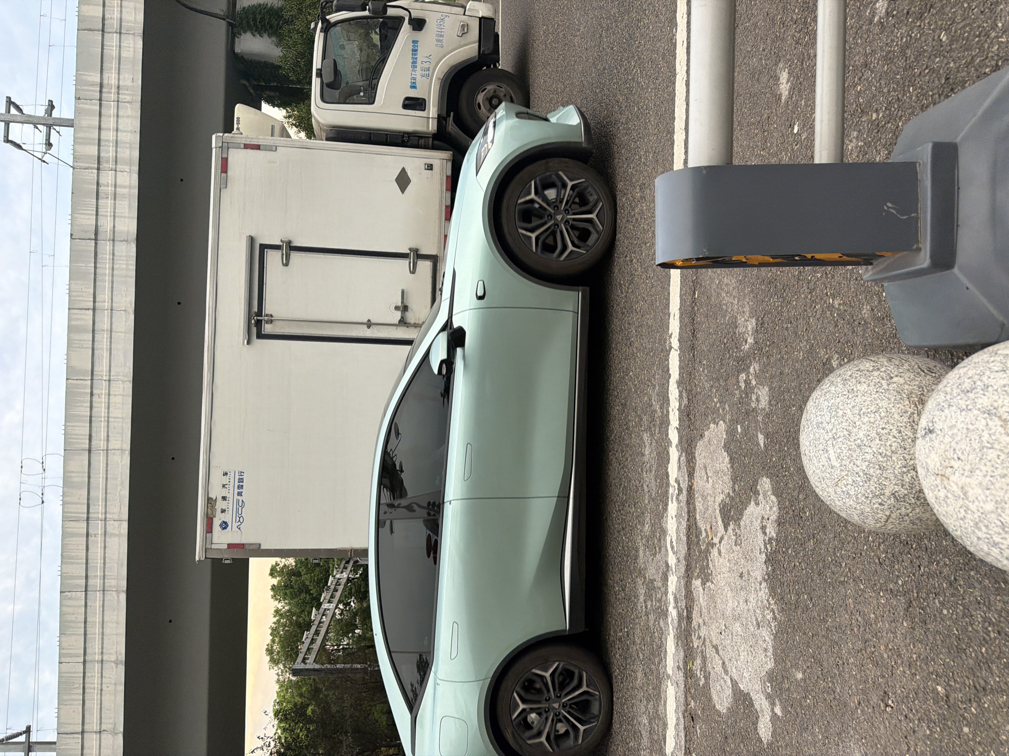 This is a medium shot, eye-level, outdoor photograph taken on a street in Jiangjin District, Chongqing, China. The image features a light green sedan car driving on an asphalt road with white lane markings. Behind the sedan, a white box truck is parked. The truck has some Chinese characters and logos on its side. The background shows a concrete overpass, trees, and power lines against a cloudy sky. In the foreground, two large, round, grey, speckled decorative stones are visible, along with a grey traffic bollard with yellow reflective elements. The scene appears to be captured during daylight.