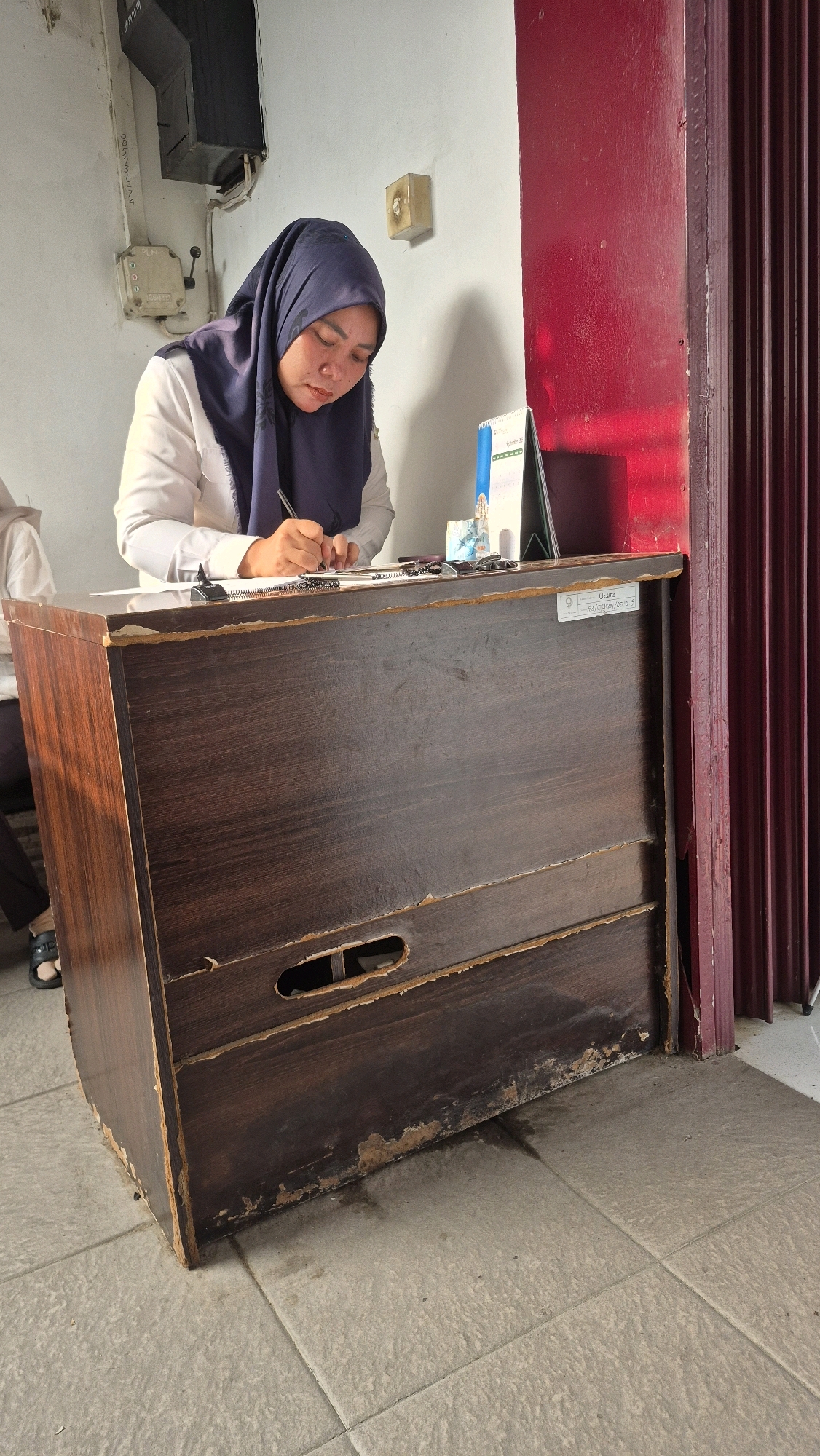 A woman wearing a dark blue hijab and a white shirt is seated at a dark brown wooden desk, writing with a black pen. She is looking down at a document on the desk. To her right, there is a desk calendar and a dark red, corrugated metal door frame. The desk itself is worn and shows signs of damage, with peeling laminate along the bottom edge. The floor is tiled with light gray square tiles, and there is some wetness on the floor in front of the desk. In the background, on the white wall, there is a black electrical box with a switch and some writing on it. Another person's arm and shoulder are visible to the left of the woman. The overall impression is of a modest, functional office or reception area. Given the context of the desk name tag which reads "Utama - S1/081/20/-6.15", this scene likely takes place in the city of Jambi, Indonesia, possibly at a government office or public service building, as "Utama" often refers to a primary or main office in Indonesia, and the date format is consistent with Indonesian date conventions.