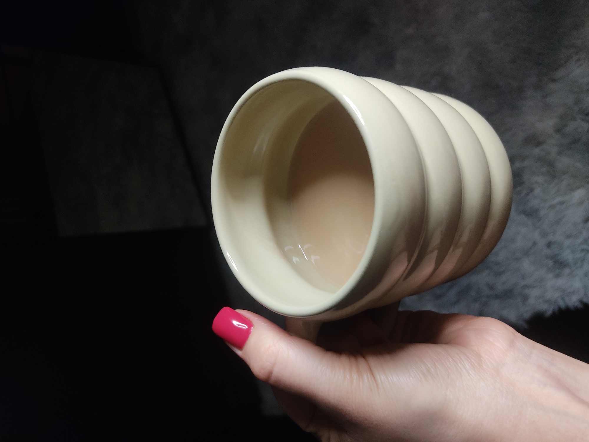 A human hand with pink fingernails holds a ribbed, beige-colored mug filled with a light brown liquid. The background is dark, with fuzzy material visible on the right side. The location is Unknown, Unknown.