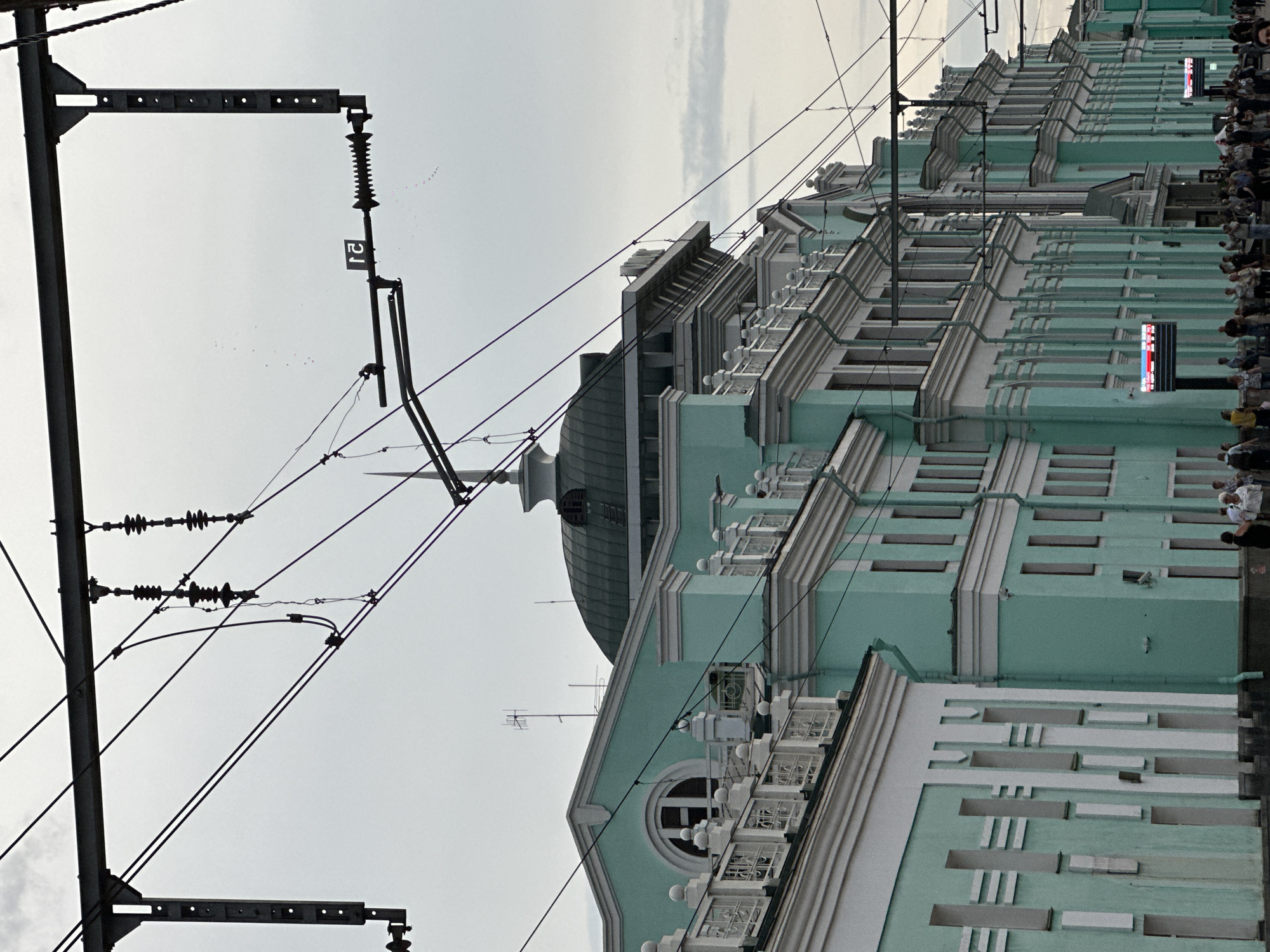 The image captures the facade of the Omsk-Passazhirskiy railway station in Omsk, Russia. The building exhibits a distinctive light turquoise and white color scheme, with ornate white decorative elements along the roofline and around the windows. Several arched windows are visible, including a prominent one on the left side of the building. The roof features a large, dark green domed structure. Overhead, a complex network of trolley wires and supporting structures is visible against an overcast sky. In the foreground, a crowd of people is gathered, some walking and others standing, suggesting activity at the station. Digital display boards with information are present on the facade. The overall scene depicts a functional urban environment centered around a major transportation hub.