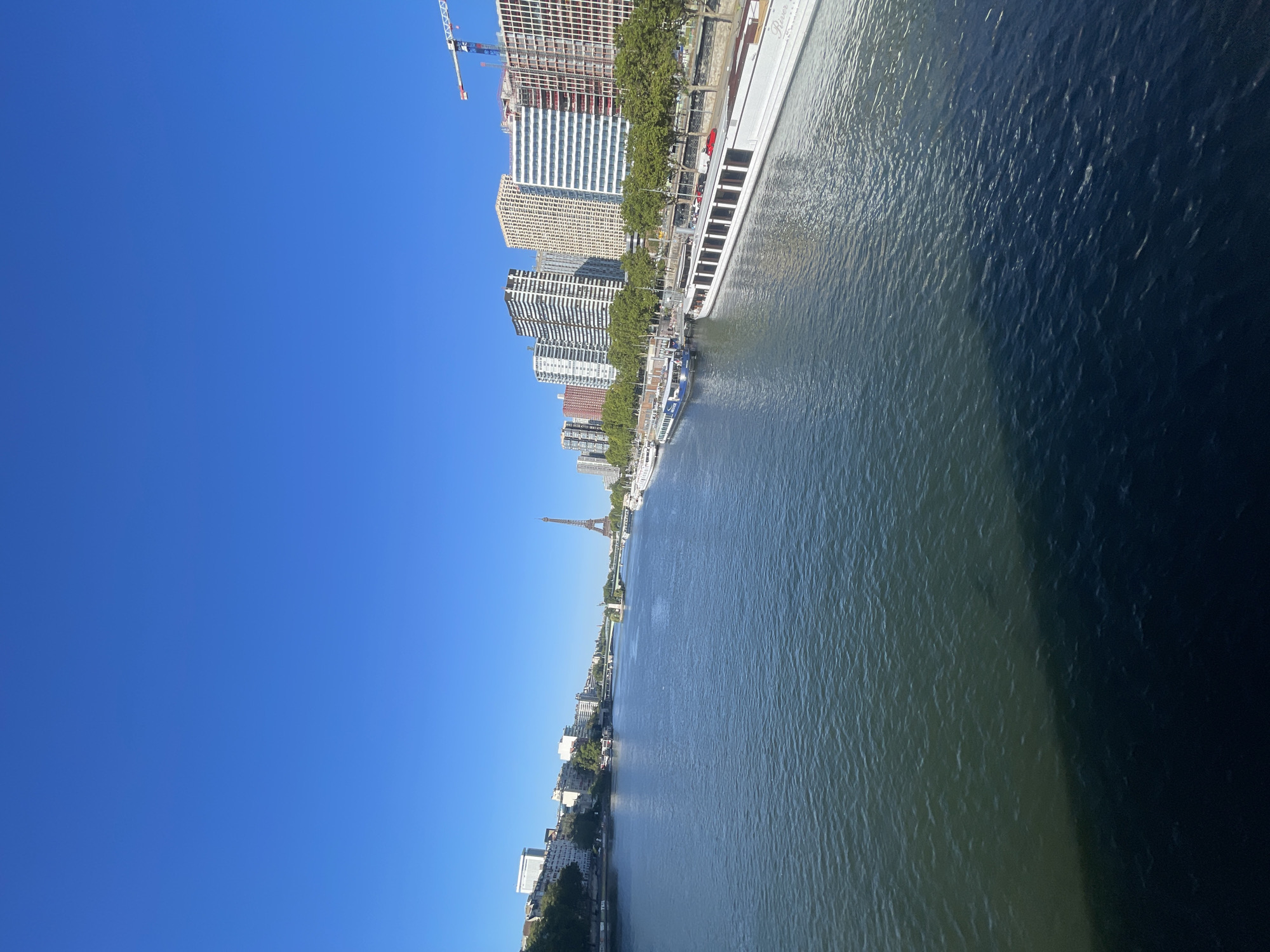 This is a photograph of the Seine River in Paris, France, on a clear, sunny day. The Eiffel Tower is visible in the distance, along with several bridges spanning the river. Along the riverbanks, there are buildings, trees, and several boats, including what appears to be a tour boat. The water is calm with some ripples, and the sky is a vibrant blue. A construction crane is visible on the right side of the image, indicating ongoing development.