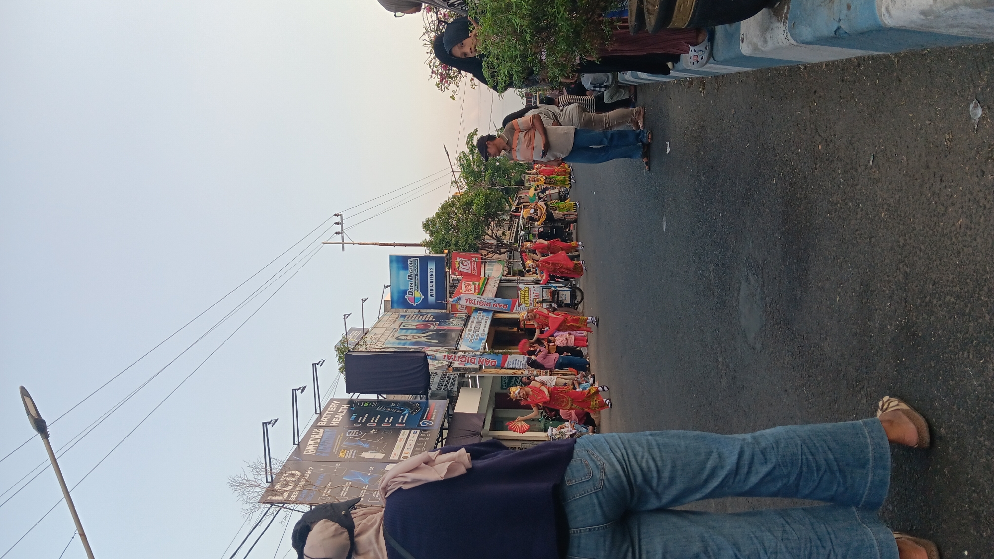A vertical, medium-wide shot captures a street scene at dusk in Tanjung Anom, Indonesia, with a cultural performance in progress. The foreground features the lower body of a person wearing wide-leg blue jeans and brown and white patterned slip-on shoes, viewed from the rear. To the right, a group of people stands along a street curb with a green bush in a black planter. The street itself is asphalt, with a raised curb marked by blue and white stripes. In the mid-ground, several children in traditional red and gold costumes, some holding red fans, are performing a dance. They are accompanied by adults, also in traditional attire. Behind the performers, a busy street scene unfolds with shops and storefronts lining the road, adorned with various signs and advertisements. A few motorcycles are parked on the side of the road. The sky above is a pale, hazy blue, suggesting the late hour. The overall impression is of a community event taking place in a lively urban environment.