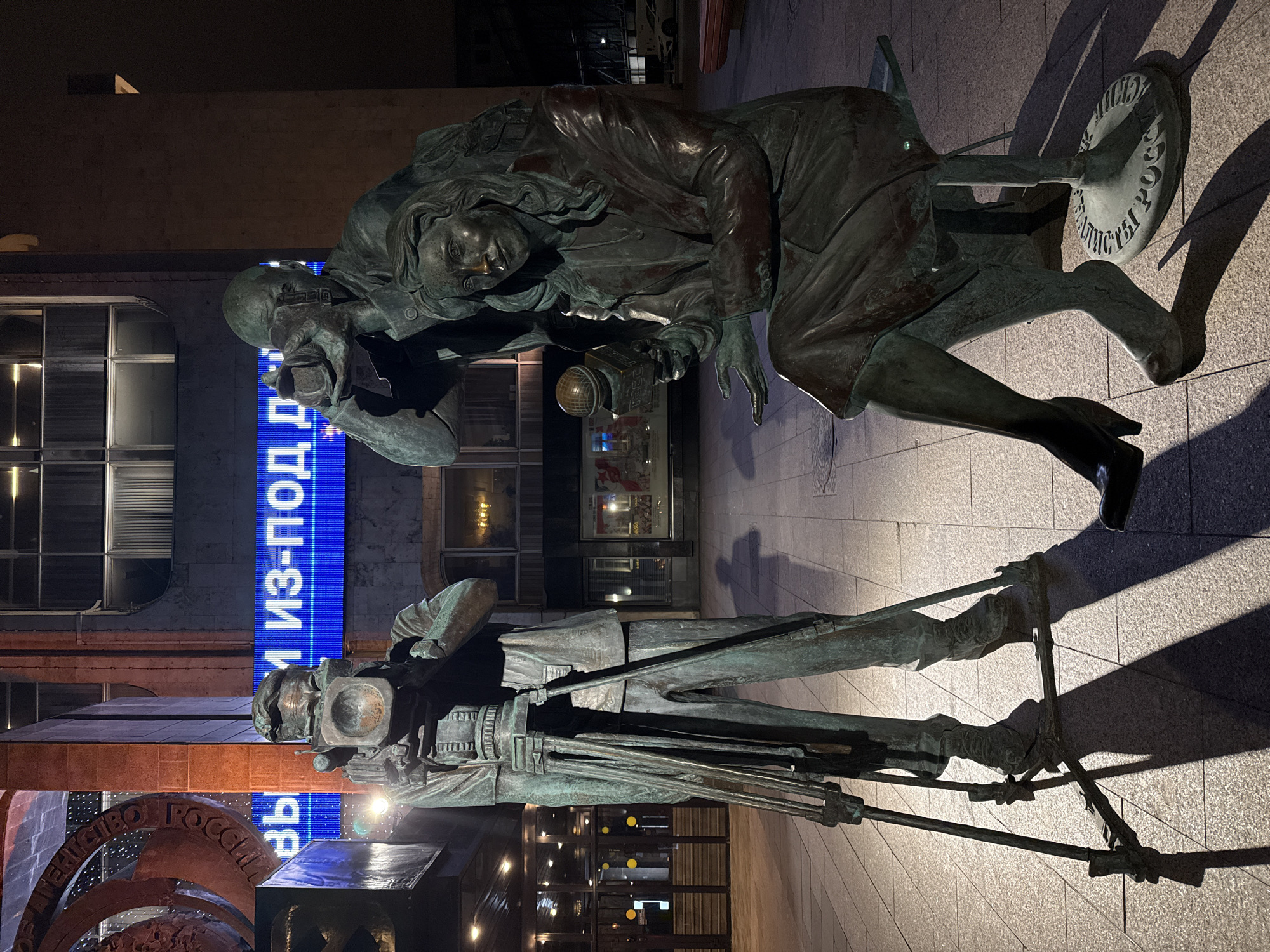 The image shows a bronze monument depicting a photographer and a female reporter in Moscow, Russia. The photographer stands with a large format camera mounted on a tripod. The female reporter kneels beside him, holding a microphone. They are positioned on a paved surface, with modern buildings and illuminated signage in the background. The lighting suggests it is nighttime.