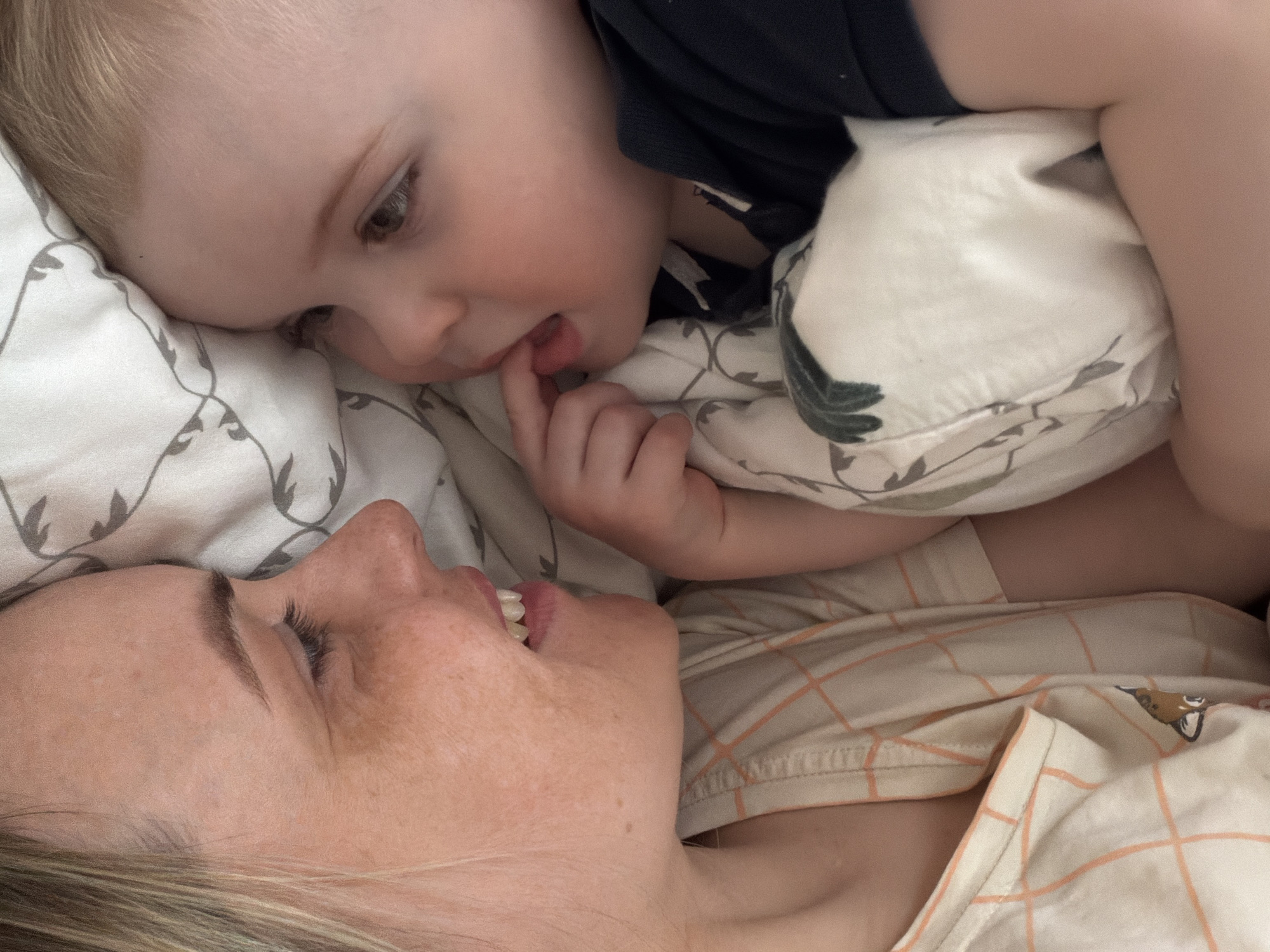 A close-up shot from an oblique angle captures a mother and her child lying on a bed with patterned bedding. The mother's face is prominently visible, showing freckles and blonde hair. She is looking upwards and slightly to her left, mouth open slightly. The child, possibly a toddler, is positioned above her, also gazing in her direction. The child has their fingers near their mouth. Both appear to be in a comfortable indoor setting. The location is not discernible from the image.