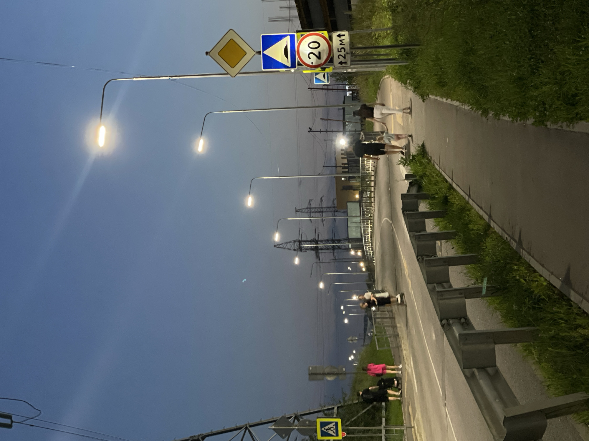 The image captures a street scene at dusk in Odintsovo, Russia. A paved pedestrian walkway runs along the right side of a road with a metal guardrail separating it from the roadway. Several streetlights are illuminated, casting light on the area.
On the walkway, a family is seen walking: an adult male, an adult female, and a young child holding hands. To their left, further down the walkway, another person is riding a scooter. On the far left of the image, near a large power line tower and a pedestrian crossing sign, a group of people are standing, one of whom is pushing a stroller.
Several traffic signs are visible: a yellow diamond-shaped sign, a blue sign with a yellow triangle indicating a speed bump, a red and white circular sign displaying "20", and another smaller blue sign with a yellow triangle. The sky is a deep blue, indicating twilight. The overall environment suggests an urban or suburban setting with ongoing construction visible in the background.
