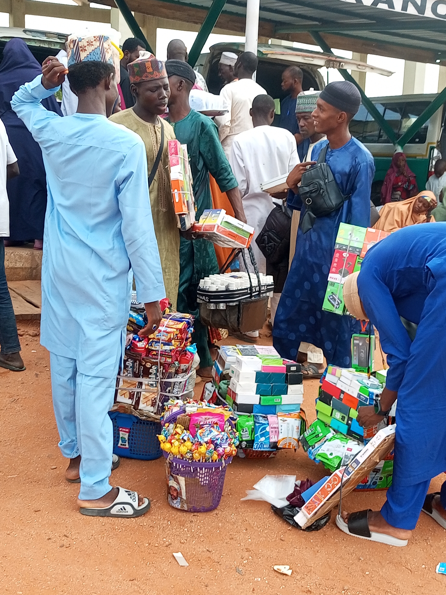 The image depicts a bustling market scene likely in Gombe, Nigeria. Several individuals, predominantly men and some women in traditional attire, are interacting around stalls laden with various goods. The foreground is dominated by a display of packaged snacks, confectionery, and what appear to be electronics or small appliances, organized in boxes and baskets.
In the center of the image, a young man wearing a light blue traditional outfit is adjusting his cap. To his right, another man in a tan-colored garment holds a stack of colorful boxes. Further to the right, a man in a deep blue outfit carries a black bag over his shoulder and is looking to his side. In the background, a bus or truck with a green and white exterior is visible, suggesting a transportation hub or a busy thoroughfare where such markets often set up. The ground is covered in reddish-brown earth, typical of many areas in Gombe. The overall atmosphere suggests a lively commercial exchange, with vendors and customers engaged in transactions.