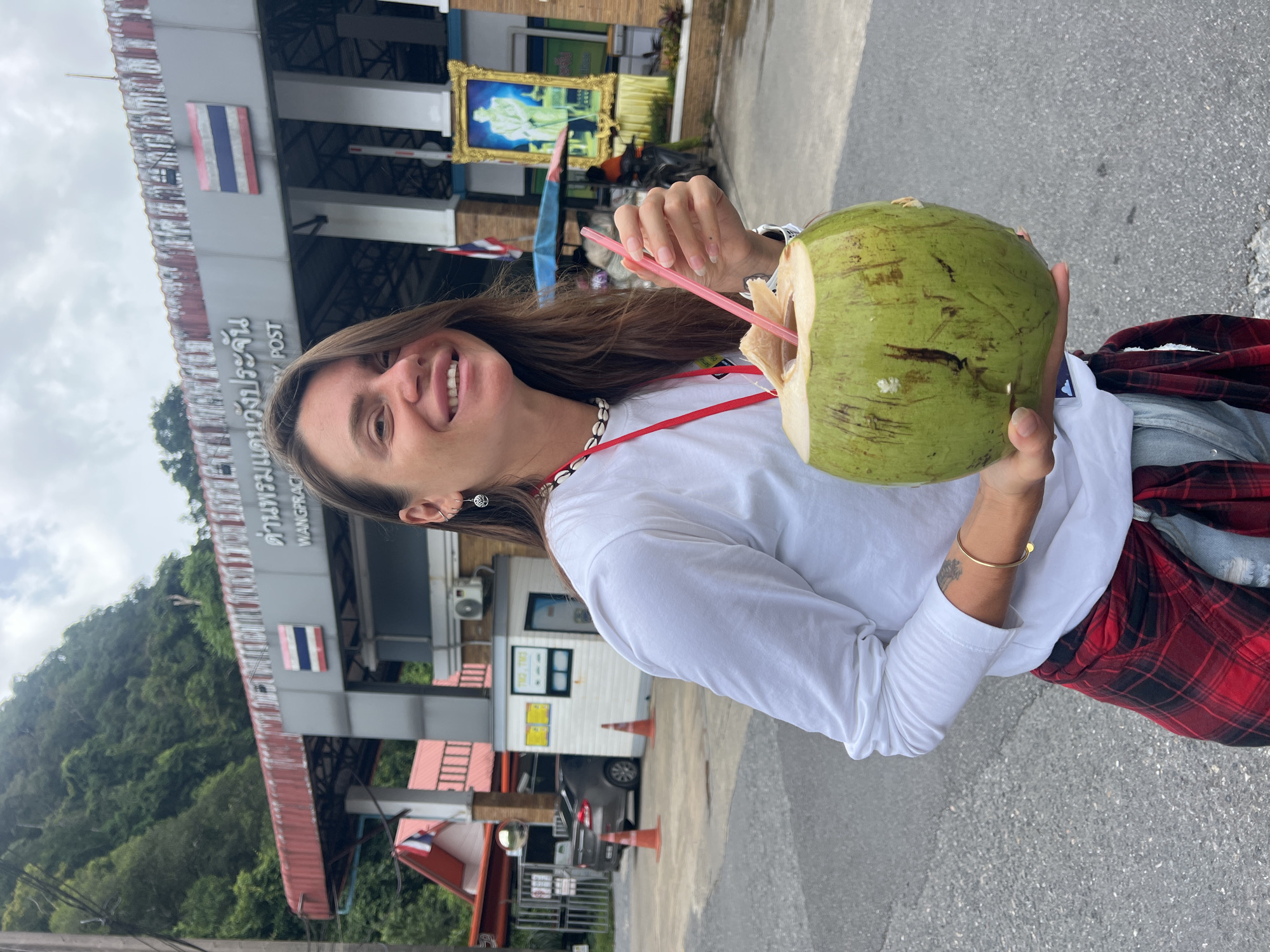 A woman with long brown hair, wearing a white long-sleeved shirt, ripped blue jeans, and a red and black plaid shirt tied around her waist, stands outdoors holding a large, green coconut with a pink straw. She is smiling and looking at the camera. Behind her is a building with Thai writing on it, identified as "WANGPRACHAN POST." The building features a Thai flag and a framed portrait of a man in uniform. The surrounding environment appears to be a street with trees and hills in the background. The scene is set in Tambon Wang Prachan, Thailand.