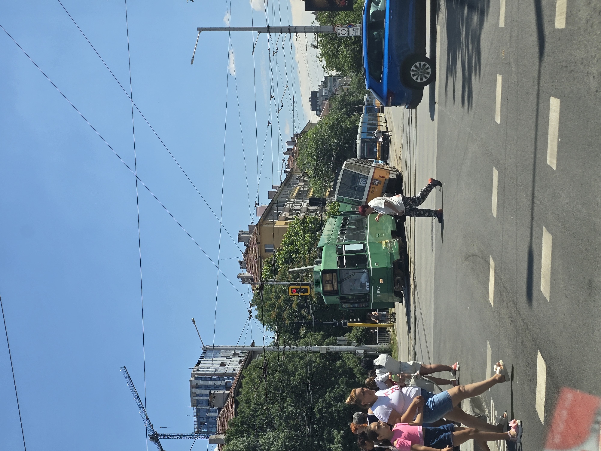 The image depicts a street scene in Sofia, Bulgaria, likely during the daytime. The foreground shows a pedestrian crossing with several people walking. Two young girls, one in a pink top and blue shorts and the other in a white top and denim shorts, are walking across the street. A woman in a white t-shirt and denim shorts is walking behind them. Further back, a person in a white shirt and patterned pants is crossing the street.
The midground is dominated by a green tram, with the number "677" visible on its front. Another tram, yellow and white, appears to be tilted at an angle behind the green tram. Several other trams are visible further down the street. There are also streetlights and overhead wires for the trams.
In the background, buildings with red-tiled roofs and a taller modern building under construction are visible. A construction crane is also present. The sky is clear blue with some clouds. The overall impression is a busy urban environment with active public transportation.