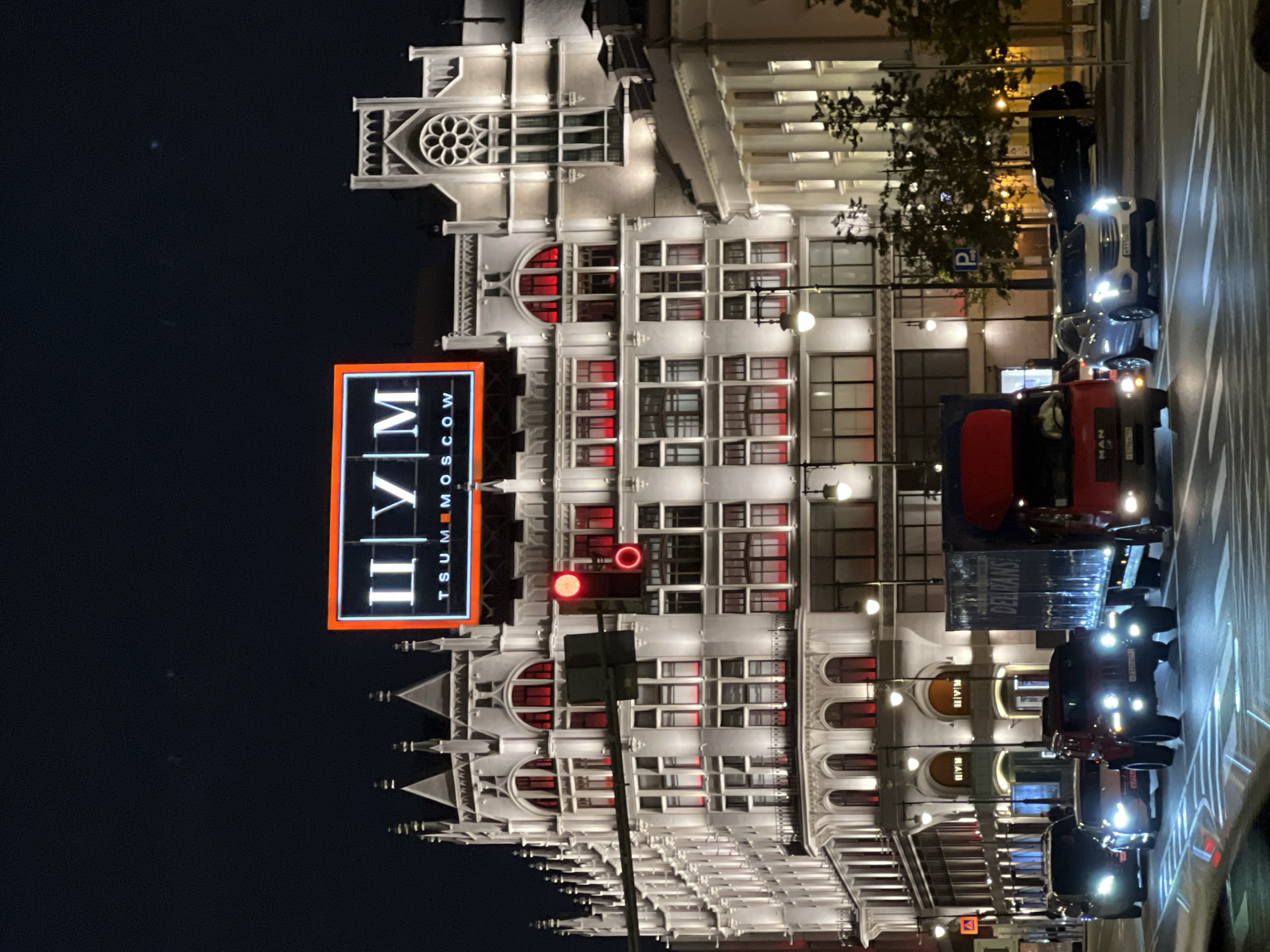 The image captures a nighttime street scene in Moscow, Russia, dominated by the illuminated facade of the TSUM department store. The architecture of TSUM is ornate, featuring white stonework, numerous windows with red illumination, and Gothic-inspired decorative elements. A large, bright sign displaying "ЦУМ" in white letters against a black background with an orange border is prominently displayed on the upper part of the building.
In the foreground, several vehicles are visible on the street, suggesting traffic. A red truck with "DELIRANS" written on its side is positioned to the right. Other cars, including a dark SUV, a red Jeep-like vehicle, and a white sedan, are in motion or stopped at an intersection, indicated by headlights and taillights. A traffic light with a red signal is visible on a pole attached to the TSUM building. Streetlights cast a warm glow on the scene, and trees with illuminated foliage line the sidewalk. The overall impression is a bustling urban environment at night, with the iconic TSUM department store as the central focal point.