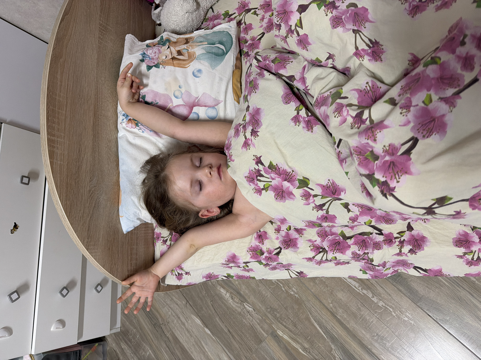The image displays a young child sleeping in a bed within a residential room in the city of Chaikovsky, Russia. The child, appearing female with brown hair, lies on her back with eyes closed and arms extended upwards and slightly outwards. She is covered from the chest down by a duvet featuring a pink cherry blossom pattern on a cream background. Her head rests on a white pillow decorated with a cartoon-style mermaid and bubbles. A small gold-colored hoop earring is visible in her right ear. The bed frame is light wood-toned with a curved headboard. To the immediate left of the bed, a white dresser with three visible drawers and square silver-colored handles is positioned. A small sticker, possibly depicting an animal, is affixed to the dresser or adjacent wall. The floor consists of light brown wood-look laminate planks. A grey stuffed animal is partially visible on the bed near the child's head.