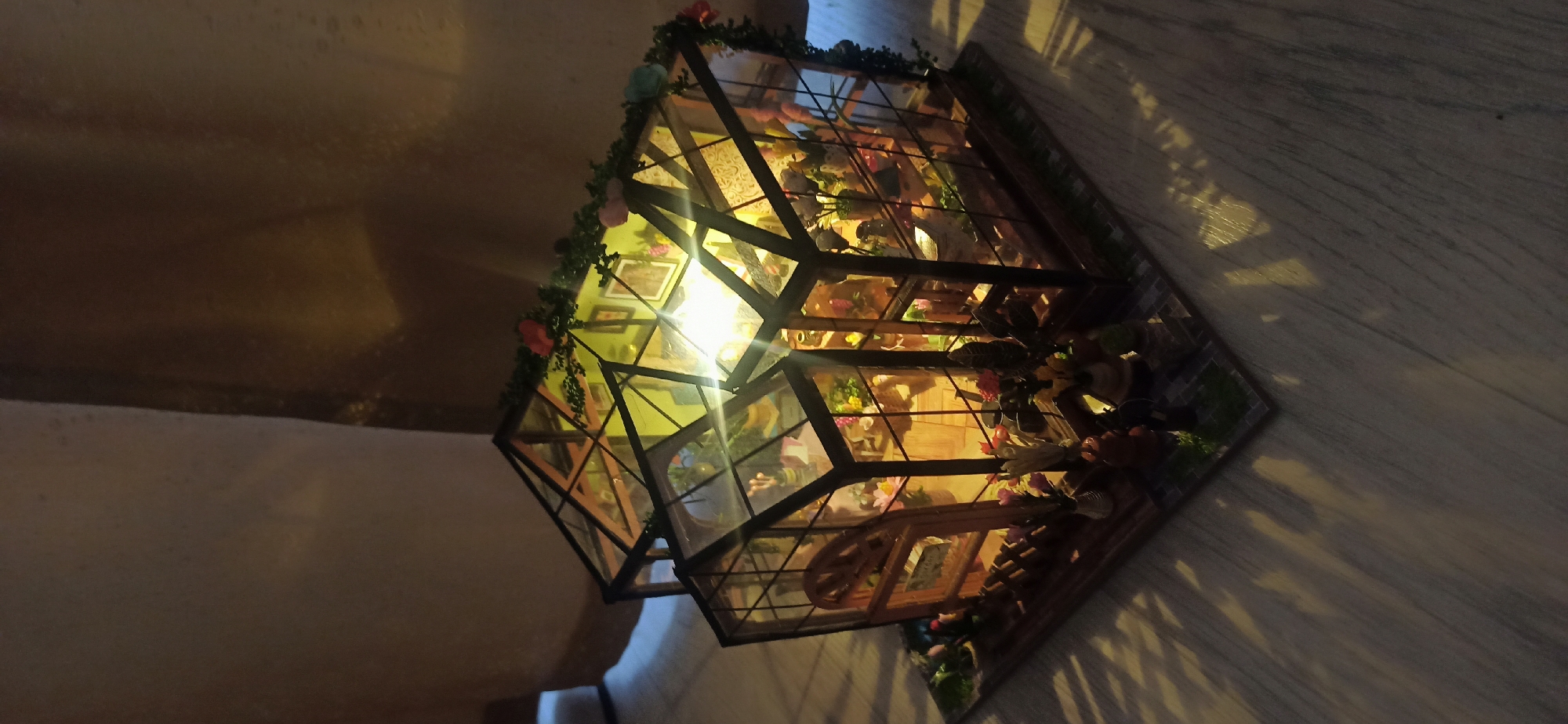 The image shows a miniature greenhouse diorama, likely a craft or hobby item. The greenhouse is made of glass panels and dark framing, adorned with small green vines and colorful faux flowers. Inside, it is illuminated by a bright light source, revealing various miniature objects such as plants, vases with flowers, and possibly small figurines. The diorama is placed on a marble-like surface, with light casting dappled shadows across it, suggesting the lighting is coming from an external source. The scene appears to be indoors, possibly in a home environment, given the flooring and ambient lighting. There is no visible text in the image. The overall impression is one of intricate detail and a charming, whimsical miniature world.