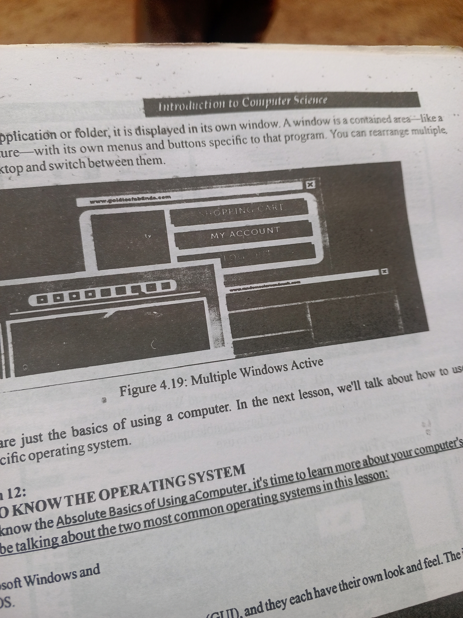 This media file shows a close-up of a page from a book, likely a textbook or manual, open on a surface. The page features text and an illustration. The text includes the heading "Introduction to Computer Science" and a paragraph describing application windows. Below this, there is a black and white illustration of multiple computer windows active on a screen, with headings like "SHOPPING CART" and "MY ACCOUNT." Further down the page, there is a caption "Figure 4.19: Multiple Windows Active" and more text that appears to be part of a lesson, including the phrase "TO KNOW THE OPERATING SYSTEM" and a sentence about learning about computer's operating systems. The text also mentions "Microsoft Windows and OS." The overall scene suggests an educational setting. There are no people visible in the image. The lighting appears to be from above, typical of indoor lighting. The visible text is in English. There are no specific location cues within the text or image that would indicate Jalingo, Nigeria, or any particular time of day or weather conditions.