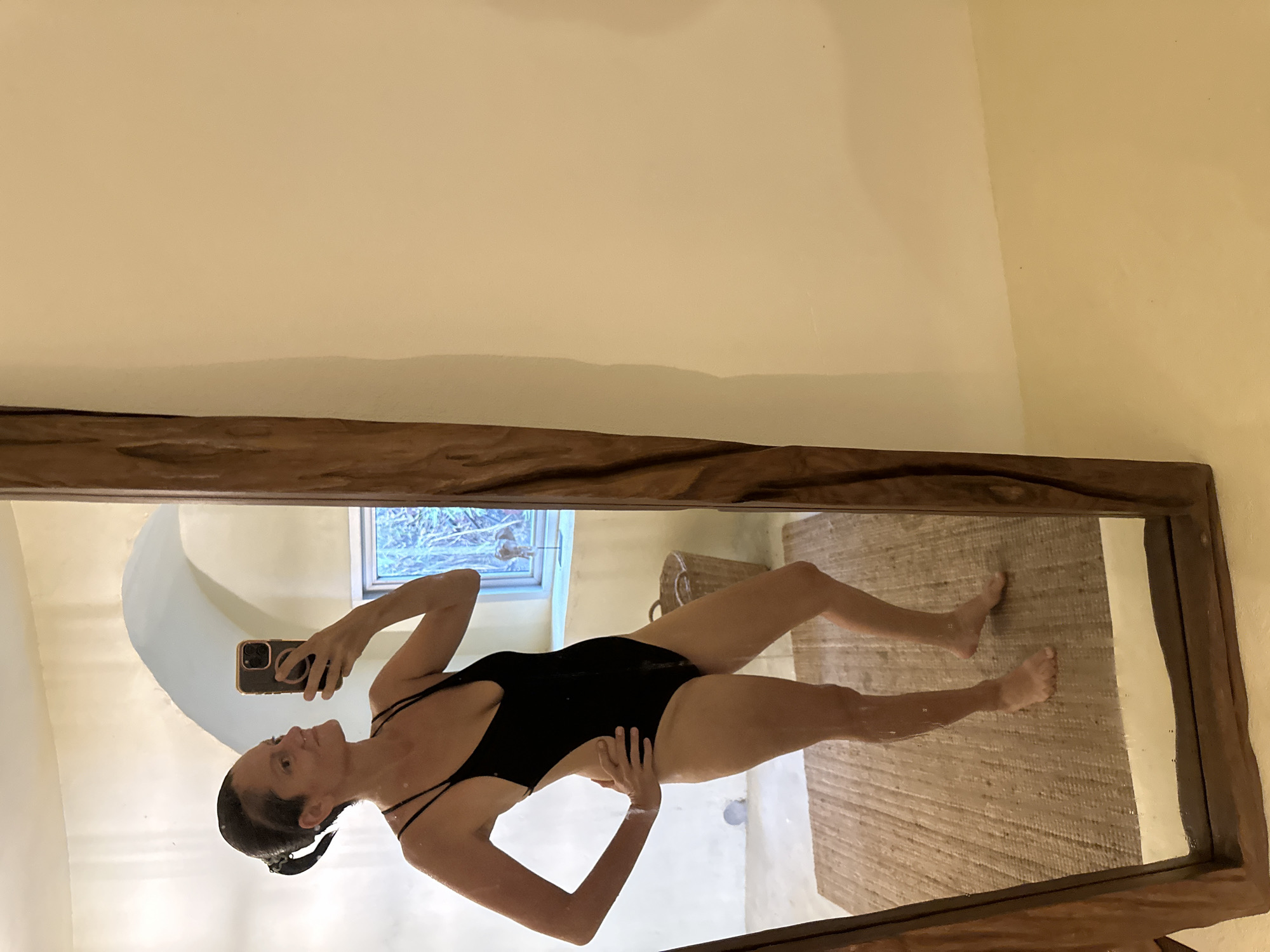 A woman is captured in a full-body mirror selfie, appearing to stand upright as reflected in the large, rectangular mirror. She wears a black one-piece swimsuit with thin straps and has dark hair pulled back. She holds a smartphone with a dark case in her left hand, looking towards it to take the picture, while her right hand rests on her hip.
The mirror is framed by a thick, rustic wooden border. The reflection reveals a bright, light-colored room with smooth walls that show some curvature, suggesting a distinct architectural style. On the floor, a textured, woven mat or rug is visible. In the background of the reflection, a window looks out onto lush green foliage, indicating an outdoor natural setting, possibly tropical. A wicker basket is also visible in the reflection, sitting on the floor.
The scene appears to be set indoors during the daytime, with soft lighting illuminating the space. The overall impression is one of relaxation in a resort-like or natural environment. The location context provided is ตำบลเกาะพะงัน, Thailand, which aligns with the tropical surroundings suggested by the window view and rustic decor. No visible text is present in the image.
