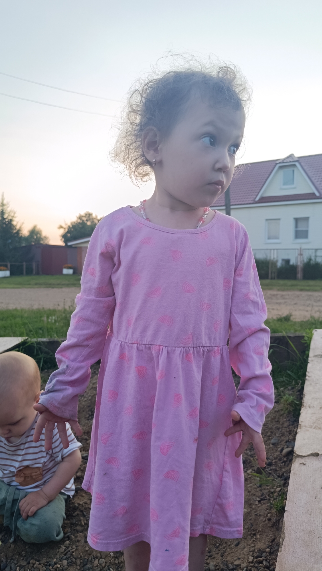 A young Caucasian girl with curly brown hair and light skin stands in the foreground, facing right and looking off-camera. She is wearing a pink long-sleeved dress with a pattern of pink semicircles. A beaded necklace with pink and clear beads is visible around her neck. Her left ear has a small hoop earring. Her hands are open and slightly raised. To her left, a much younger Caucasian child, likely a boy, is kneeling and looking down, wearing a striped t-shirt and green pants. They are positioned in what appears to be an outdoor setting with gravel and dirt on the ground, and some green plants. In the background, there is a white house with a red roof and dormer windows, along with trees and power lines against a pale sky, suggesting a rural or suburban environment. The lighting suggests it is either early morning or late afternoon. No specific identifiable landmarks within the city of Karsha, Russia, are visible.