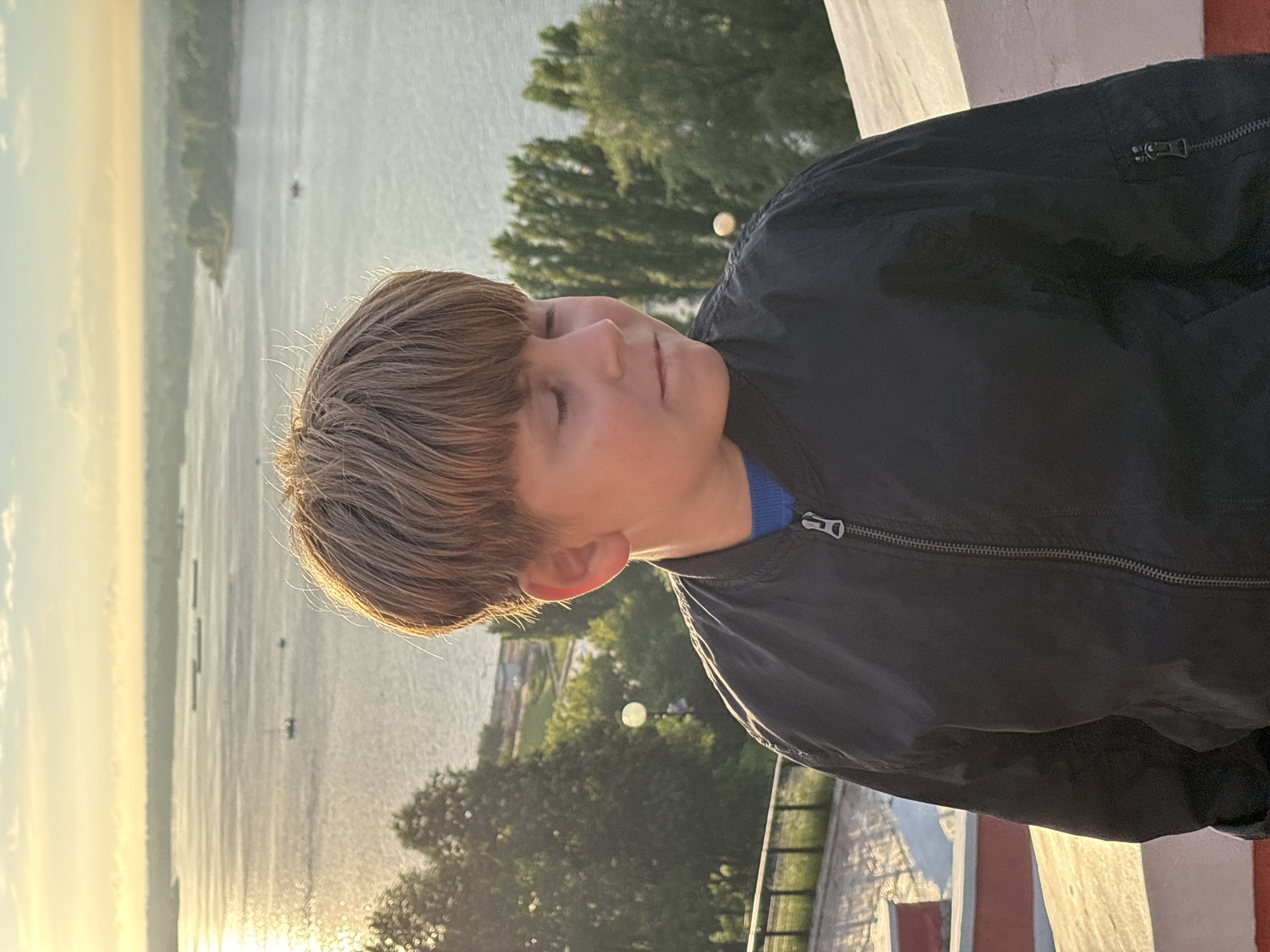 A young boy with short, tousled brown hair is pictured in a close-up, profile shot, with his eyes closed and head tilted back. He is wearing a dark blue jacket over a bright blue t-shirt. The jacket has a zipper that is partially open, revealing the t-shirt underneath. The boy's skin appears flushed, and his hair is lit by the warm glow of the sun.
The background is dominated by a body of water, likely a river, reflecting the soft, golden light of either sunrise or sunset. Distant trees and structures are visible on the far bank, suggesting an outdoor setting. The overall mood is serene and peaceful, with the boy appearing to be enjoying a moment of quiet contemplation or relaxation. The time of day is evident from the warm, low-angled sunlight casting long shadows and creating a gentle illumination.
The image appears to be taken from an elevated position, possibly a balcony or a hill, overlooking the water. In the foreground, a white railing and a red structure are partially visible, adding a sense of depth and context to the scene. The water itself has a slightly hazy quality, typical of either dawn or dusk.