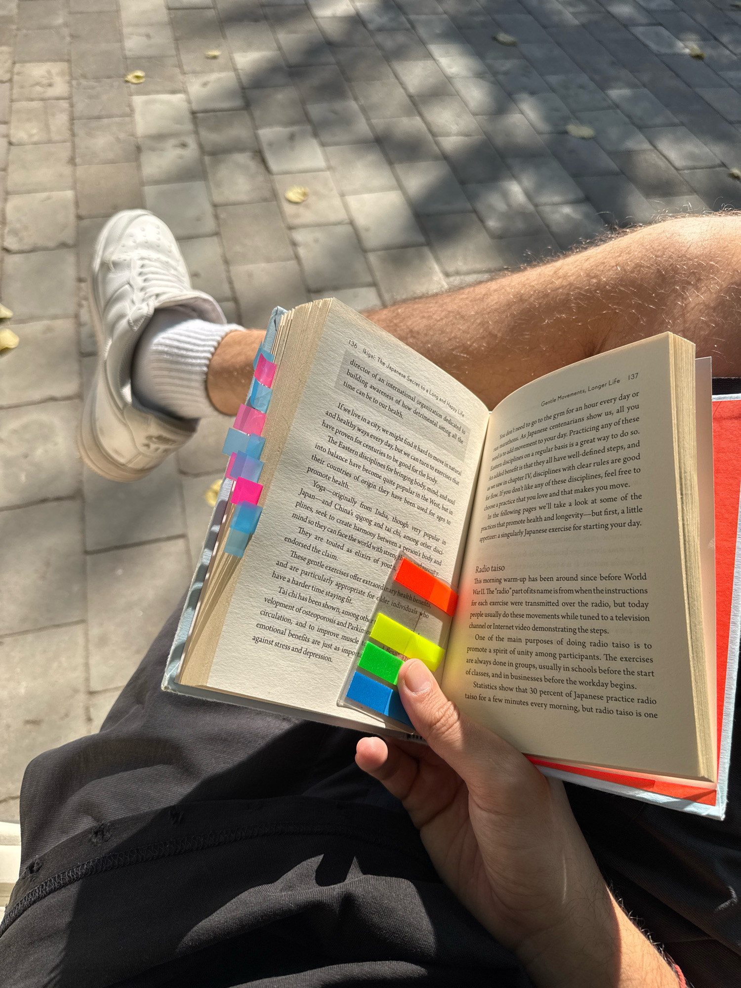 An overhead, eye-level shot shows a person reading a book outdoors, with the lower half of their body and legs visible. The person is wearing white socks and white sneakers. The book has colorful sticky tabs on the side. The ground appears to be paved with light-colored square tiles, and there is some dappled sunlight on the ground. The overall scene is casual and relaxed. The image does not contain any information about the city of Moscow, Russia.