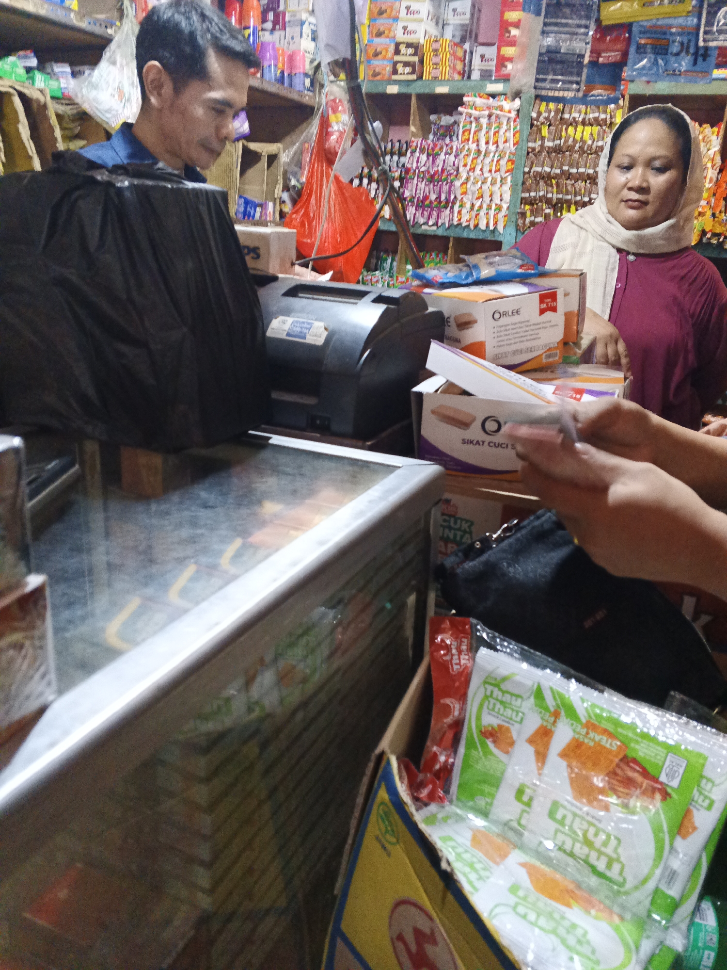 The image depicts a scene inside a small convenience store, likely a warung, in Warungkiara, Indonesia. Two individuals are visible. On the left, a man with short dark hair and a blue shirt is standing behind a counter, partially obscured by a large black plastic bag that covers a piece of equipment, possibly a receipt printer or cash register. He is looking down. On the right, a woman wearing a light-colored hijab and a dark pink or maroon shirt is standing behind the counter. Her hands are visible, holding a small plastic bag containing what appear to be monetary bills. She is looking towards the viewer's left, possibly interacting with a customer or employee not fully visible.
The environment is cluttered with merchandise stacked on shelves in the background, typical of a small Indonesian warung. Items include various packaged goods such as snacks, toiletries, and other household items. A glass-topped display case is in the foreground, reflecting the interior of the store. In a cardboard box in the lower right of the frame, several packages of snacks with green and white packaging are visible. The overall impression is that of a busy, functional retail space in a local Indonesian community.