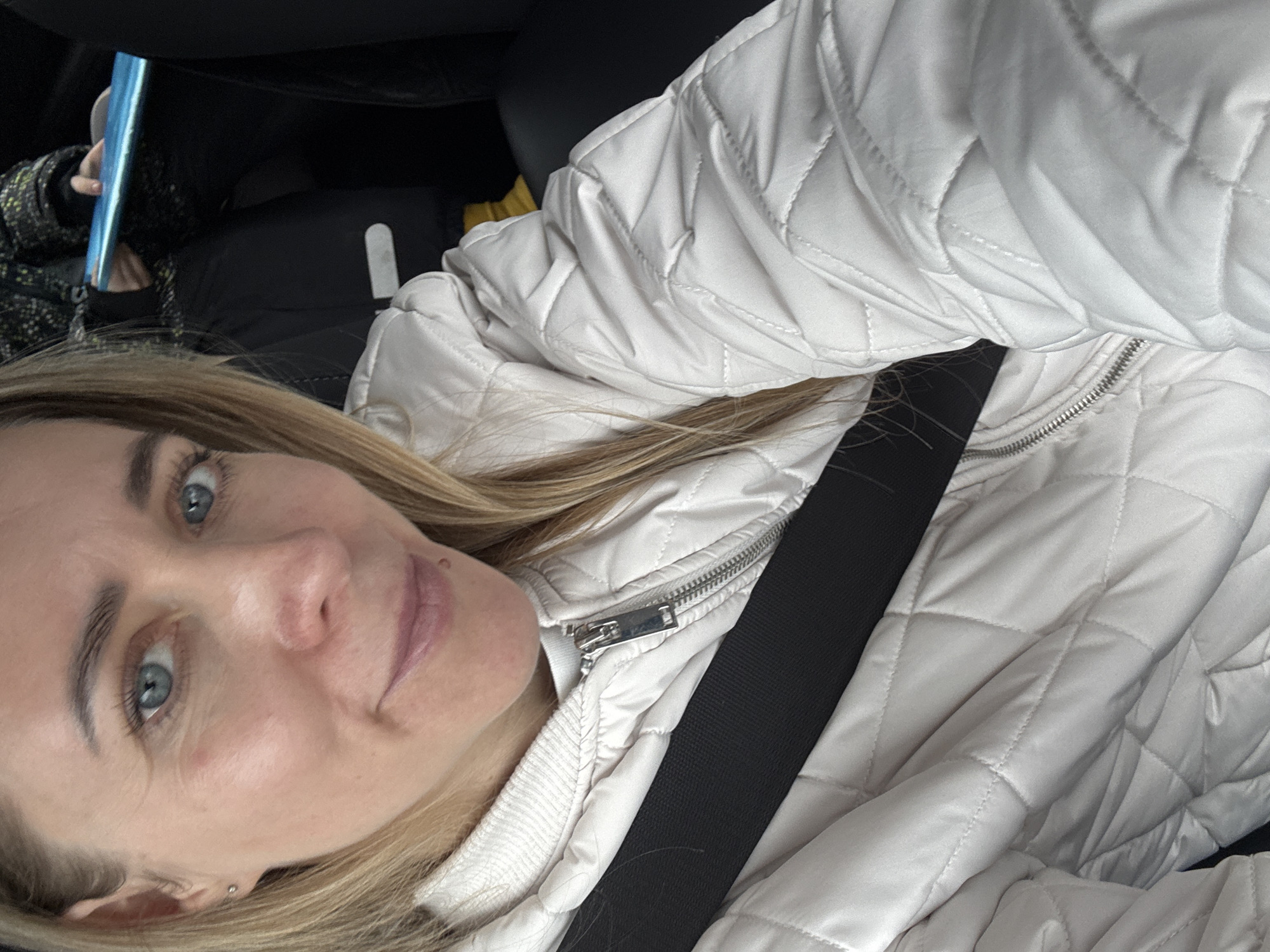 A person is wearing a white, quilted jacket and a black seatbelt. The location is not identifiable as Chaikovsky, Russia. A hand holds a blue object in the background.