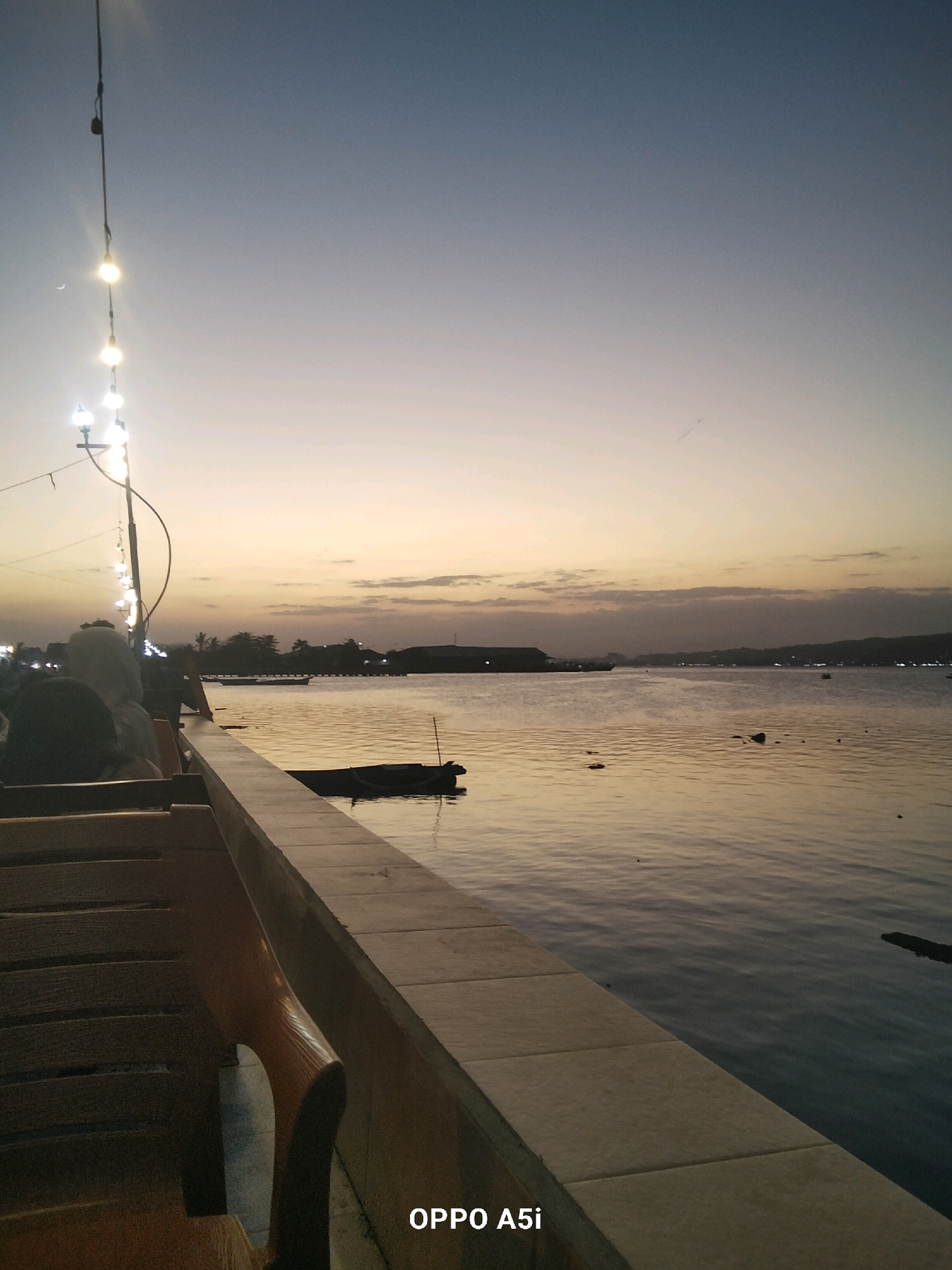 The image depicts a tranquil sunset scene over the waters of Kendari Bay, Indonesia. In the foreground, wooden benches are arranged along a stone-tiled walkway, suggesting a seating area at a waterfront establishment. A string of illuminated bulbs hangs from a pole on the left side of the frame, casting a warm glow. The calm water of the bay reflects the soft hues of the twilight sky, with a few small boats visible in the distance. The horizon is marked by a silhouette of buildings and palm trees, with the soft glow of lights indicating the presence of a populated area. The overall atmosphere is serene and peaceful, capturing the beauty of a tropical evening.