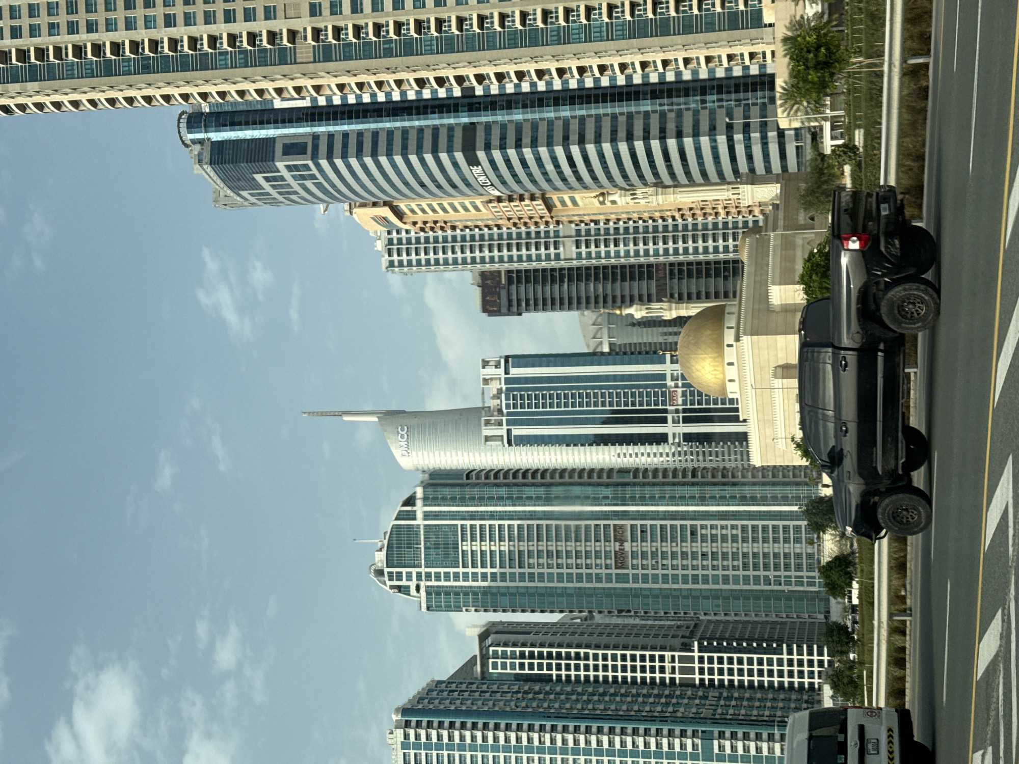 The image captures a modern urban landscape in Dubai, United Arab Emirates, characterized by an array of high-rise buildings under a clear, partly cloudy sky during daytime.
In the foreground on the right, a multi-lane road is visible, marked with yellow double lines and white dashed lines. A dark-colored pickup truck, appearing to be a GMC model, is parked or driving on this road, facing away from the viewer. The front portion of a white vehicle is also seen at the bottom right. A strip of green foliage and palm trees separates the road from the towering structures beyond.
The central and left portions of the image are dominated by a dense collection of skyscrapers, showcasing diverse architectural styles. Several buildings feature glass facades, while others display intricate patterns and balconies. Prominently visible is a tall, slender silver building with the inscription "DMCC" (Dubai Multi Commodities Centre) near its apex. Directly below it, another building clearly displays "MÖVENPICK", indicating a hotel. Another curved building with a striped pattern has "BUSINESS CENTRE" written on its side. The letters "MAG" are also discernible on a building further down. Amidst the contemporary architecture, a distinctive structure with a golden dome stands out, possibly a mosque or a decorative building.
The scene depicts a bustling cityscape, emphasizing the extensive urban development and modern infrastructure typical of Dubai.