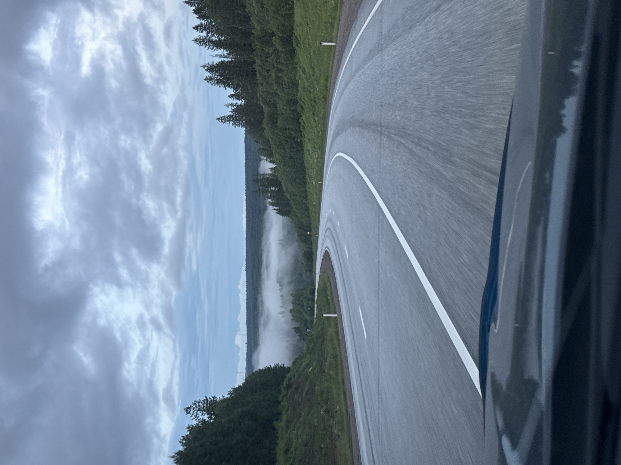 The image depicts a winding asphalt road with white dividing lines, bordered by dense green trees and grassy slopes. In the distance, a layer of fog blankets a forested valley, with a hazy sky overhead. The perspective is from inside a vehicle, with the dashboard visible in the foreground. There are no people or animals present. The scene appears to be a rural road in Div'ya, Russia, likely captured during overcast weather.