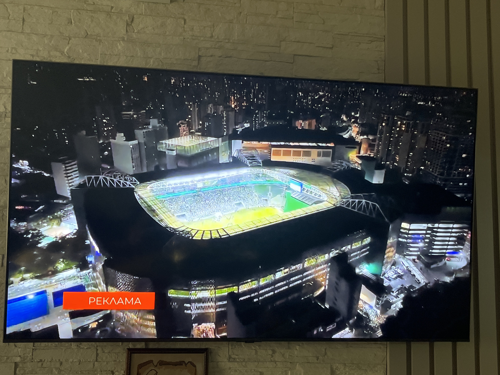 An aerial night view captures a stadium filled with spectators and illuminated by bright lights. The stadium is surrounded by a cityscape with numerous buildings, some of which are also lit up, indicating a densely populated urban area. The image displays a red rectangular banner with white Cyrillic text that reads "РЕКЛАМА", which translates to "ADVERTISEMENT" in English. This banner is superimposed over the bottom left portion of the stadium scene. The overall scene is presented on a television screen, which is mounted on a brick-like wall.