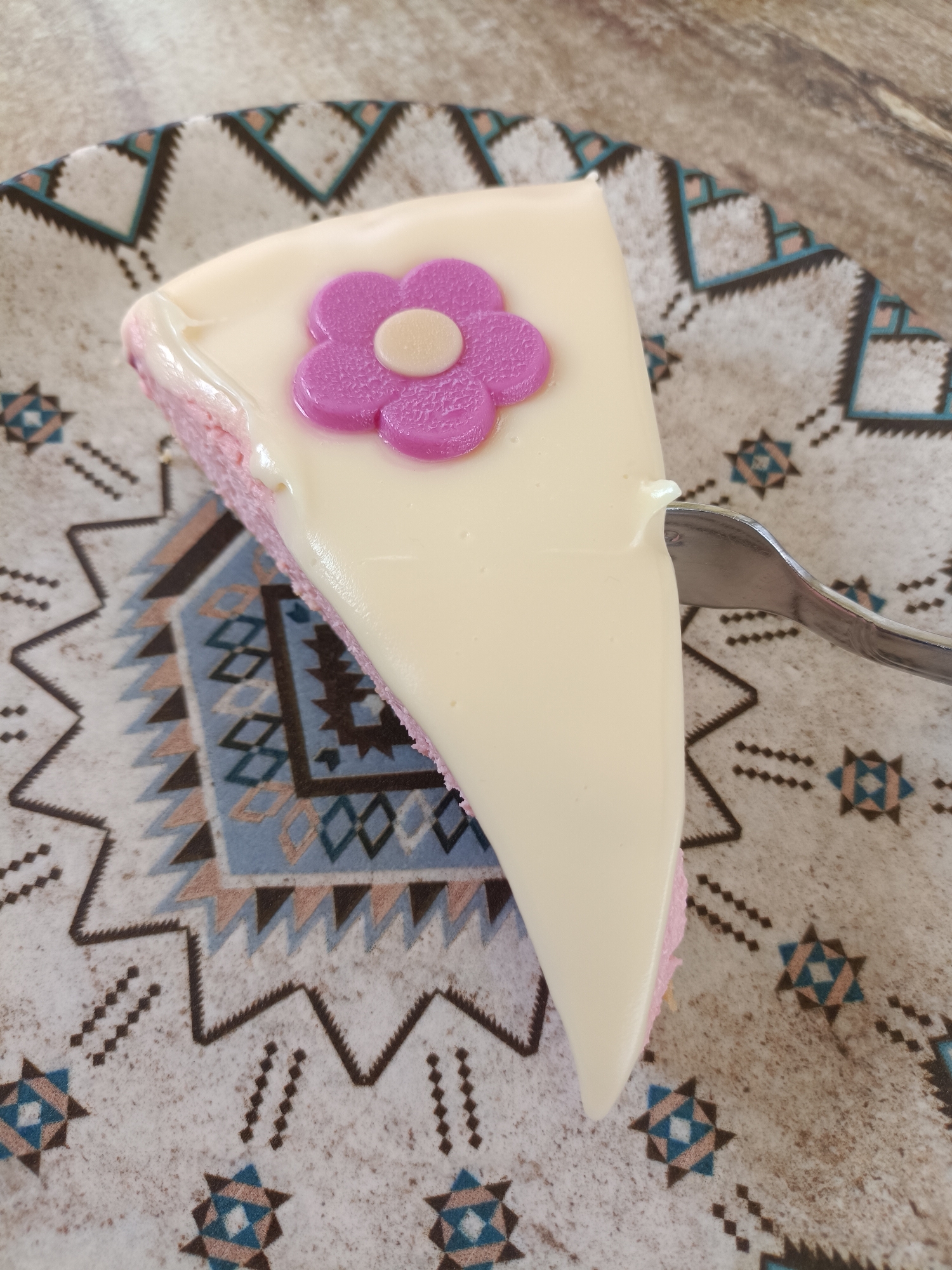 A slice of cake sits on a patterned plate, with a fork resting against its side. The cake has a light pink base, covered with a thick layer of white frosting. A purple flower with a beige center adorns the top of the slice. The plate features a geometric pattern in shades of beige, blue, and brown. The scene is likely photographed indoors. The exact location within Plovdiv, Bulgaria, cannot be determined from this image alone.