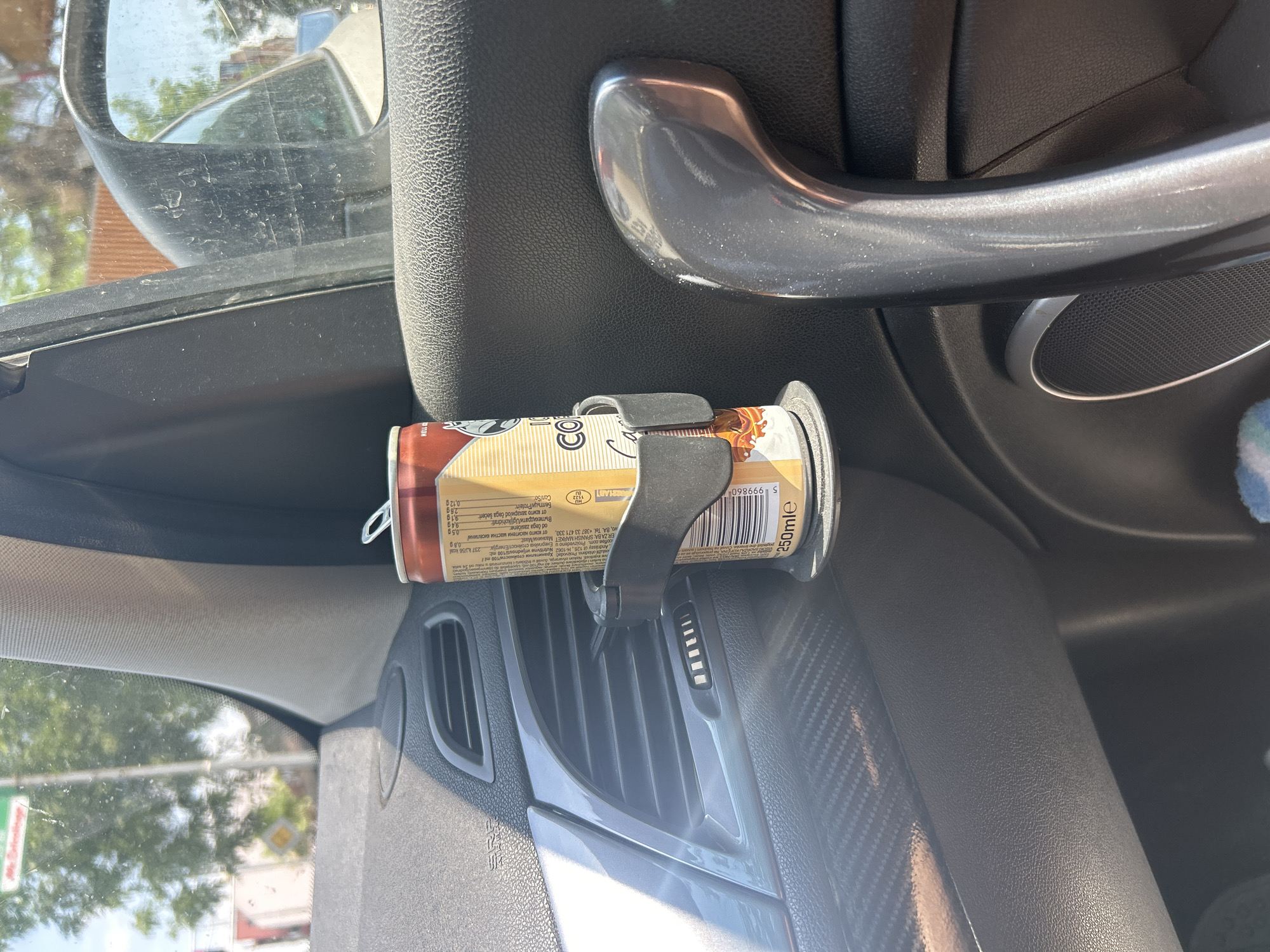 In the city of Plovdiv, Bulgaria, the interior of a car's door panel is shown. A can of "Hell Energy Coffee" is held in a car cup holder, positioned near the air vent and door handle. The can has a label with nutritional information. Sunlight streams in from the window, illuminating dust particles in the air. The background shows a blurred view of trees and buildings.