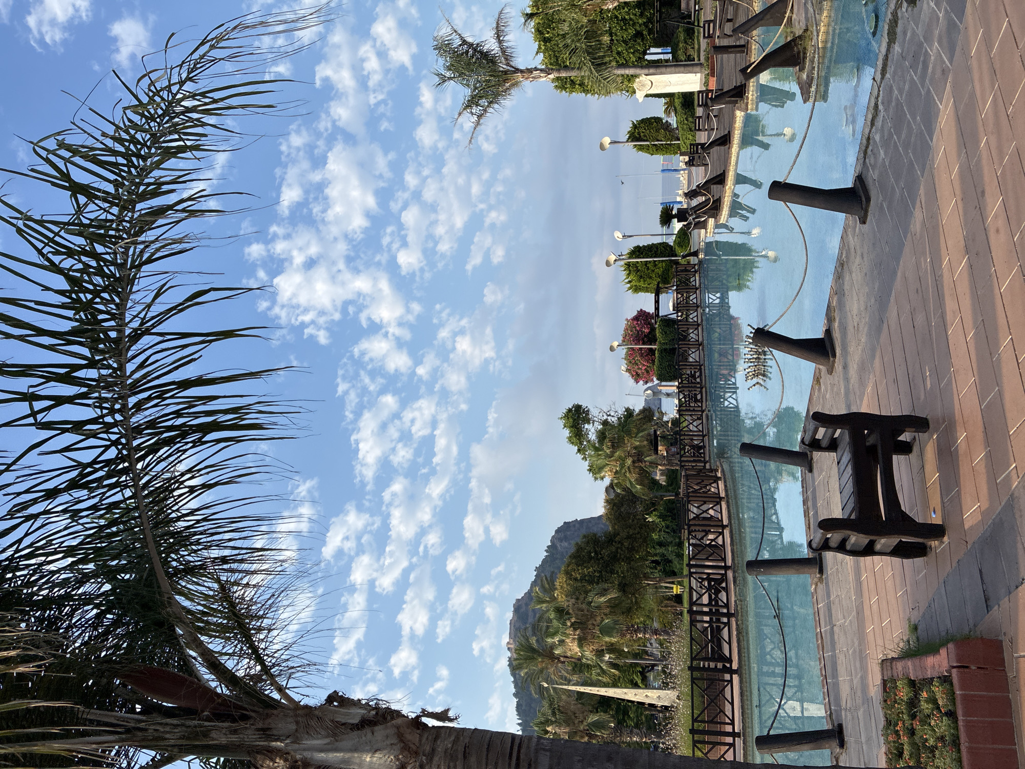 The image depicts a scenic view of Alanya, Turkey, focusing on an outdoor recreational area with a tranquil pool and lush greenery. A prominent palm tree frames the left side of the shot, its fronds reaching towards a partly cloudy blue sky. In the background, a rugged mountain with ancient ruins on its summit dominates the landscape, a signature landmark of Alanya.
The foreground features a paved walkway made of reddish-brown bricks, leading to the edge of a clear, light blue pool. Several dark wooden benches are strategically placed around the pool area, with one small, empty bench situated closest to the viewer. The pool is bordered by a low wooden fence and posts connected by rope, creating a safe and aesthetically pleasing boundary.
Beyond the pool, a variety of trees, including palm trees and dense shrubs, create a verdant backdrop. Streetlights with a modern design are interspersed among the foliage, suggesting a well-maintained public space. In the distance, the calm waters of the sea are visible, dotted with sailboats, hinting at the coastal nature of Alanya. The overall atmosphere is one of peaceful relaxation and natural beauty, capturing the essence of a Mediterranean resort town.