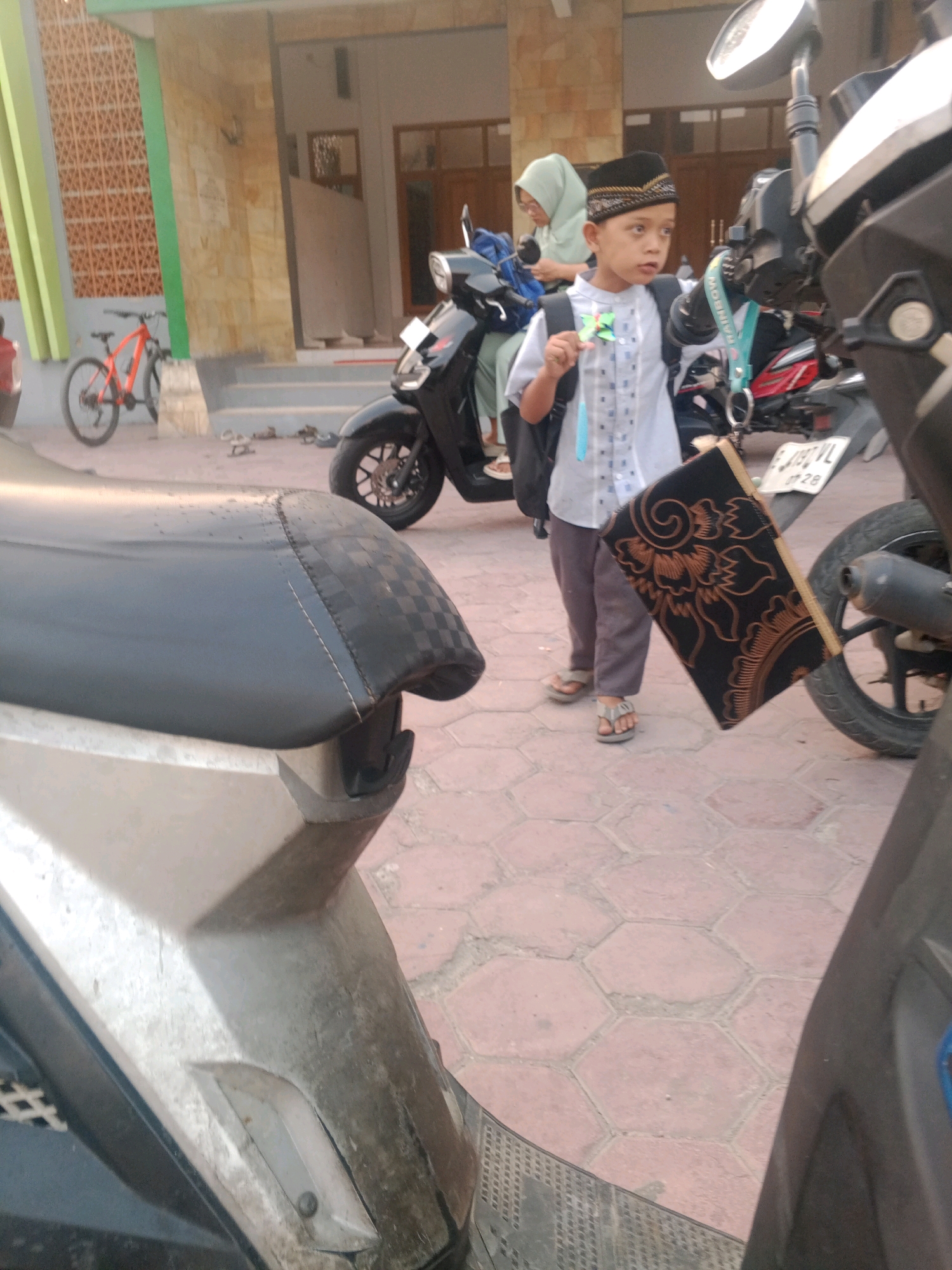 A young boy wearing a white shirt, grey pants, and a black and gold peci hat stands on a paved area outside a building, possibly a mosque or school, in Lembah, Indonesia. He is holding a small, green, star-shaped object in his right hand and has a black backpack on. A small, decorative prayer mat with an orange floral pattern is attached to a motorbike parked behind him. Another motorbike is in the foreground, partially obscuring the view. A woman wearing a light green hijab is visible in the background, near a black motorbike. An orange bicycle is parked against a wall to the left of the frame. The ground is paved with hexagonal tiles.