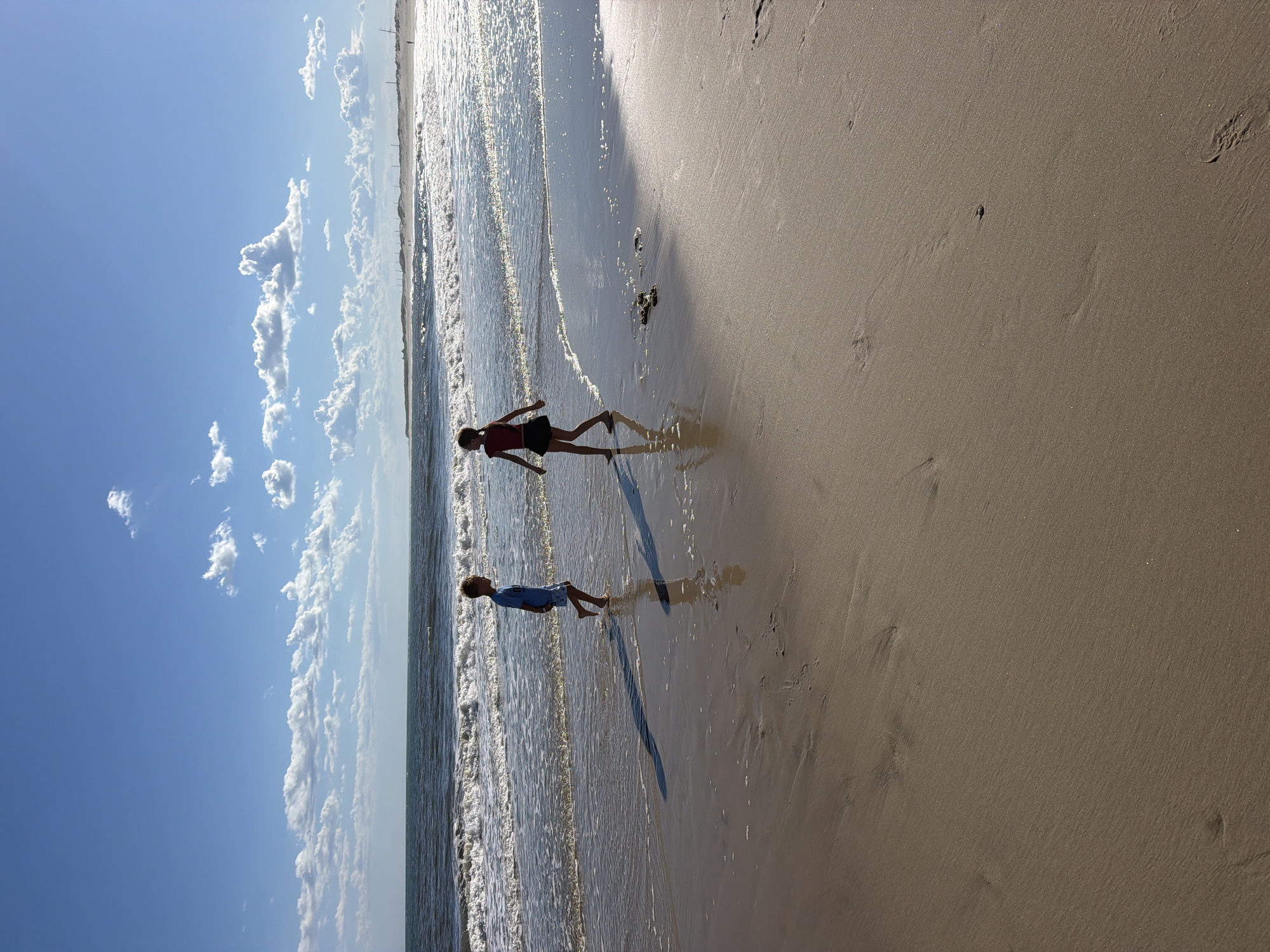 Two children, a boy and a girl, are standing on the wet sand of a beach. The boy, wearing a blue shirt and shorts, is facing away from the camera and walking towards the water. The girl, in a red top and dark skirt, is facing the boy and appears to be walking away from the water. They are silhouetted against the bright sunlight.
The ocean waves are breaking gently on the shore, with white foam visible. The sand is damp and reflects the light. In the background, the ocean stretches to the horizon under a bright blue sky with scattered white clouds. The shoreline in the distance appears to have dunes and some man-made structures, possibly wind turbines. The overall scene is peaceful and indicative of a sunny day at the beach.
The image appears to have been taken in Hampton Bays, New York, USA, a coastal town known for its beaches.