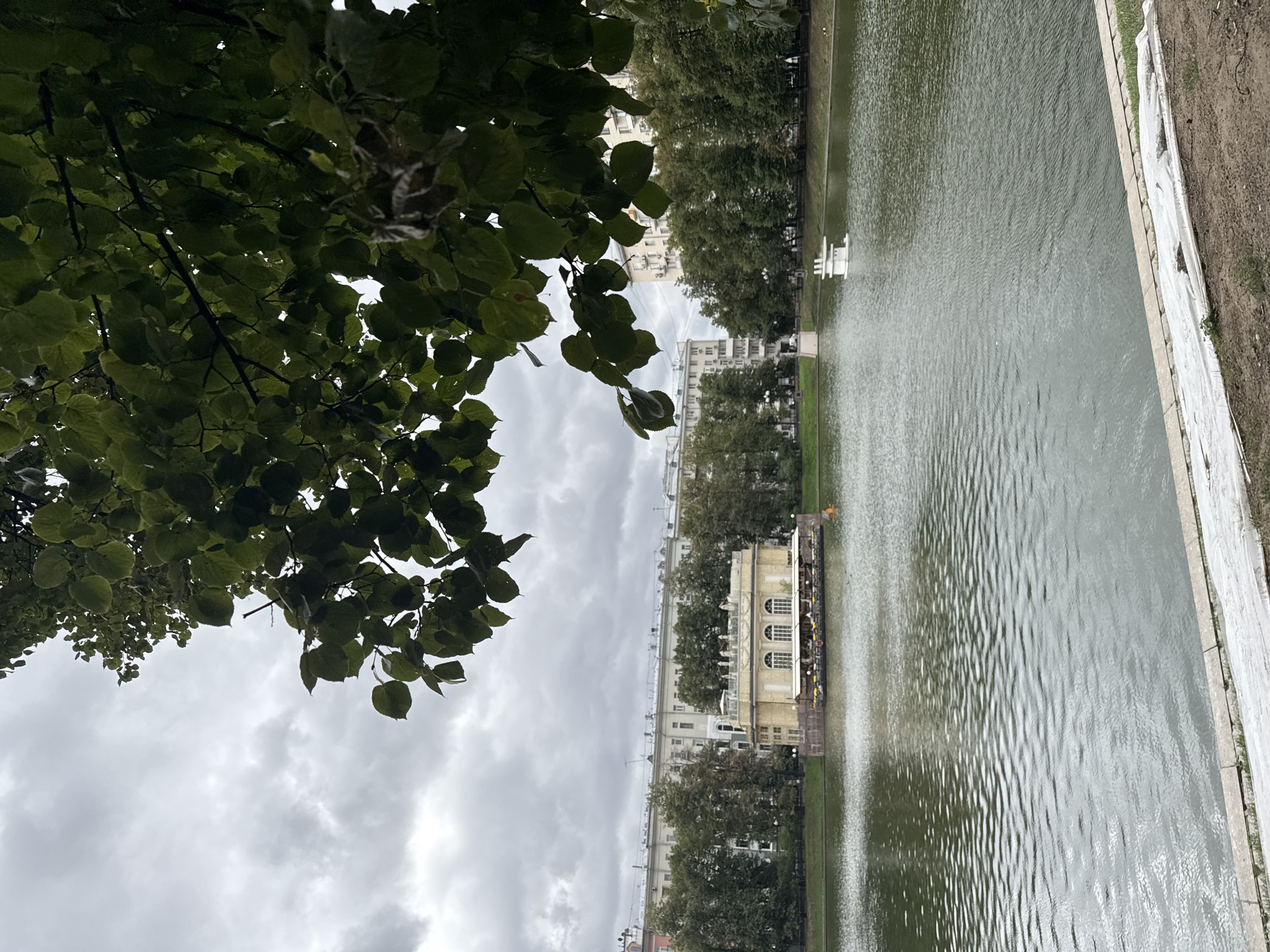 The image depicts a cloudy day in Moscow, Russia, at Patriarch's Pond. The foreground features a stone embankment with white sheeting laid over dirt. A body of water, likely the pond, dominates the middle ground, with ripples disturbing its surface. On the far side of the pond, a light-yellow building with arched windows and a white roof sits near the water's edge, appearing to be a restaurant or cafe, with tables and umbrellas visible on its patio. In the distance, several tall apartment buildings are visible, characteristic of Moscow's urban landscape, interspersed with mature trees. To the right of the yellow building, a small, white, multi-tiered structure, possibly a decorative element or part of a fountain, is situated in the pond. The sky is overcast with thick, gray clouds, contributing to the muted lighting of the scene. The branches of a large tree with green leaves hang into the frame from the top, partially obscuring the view of the sky and buildings.