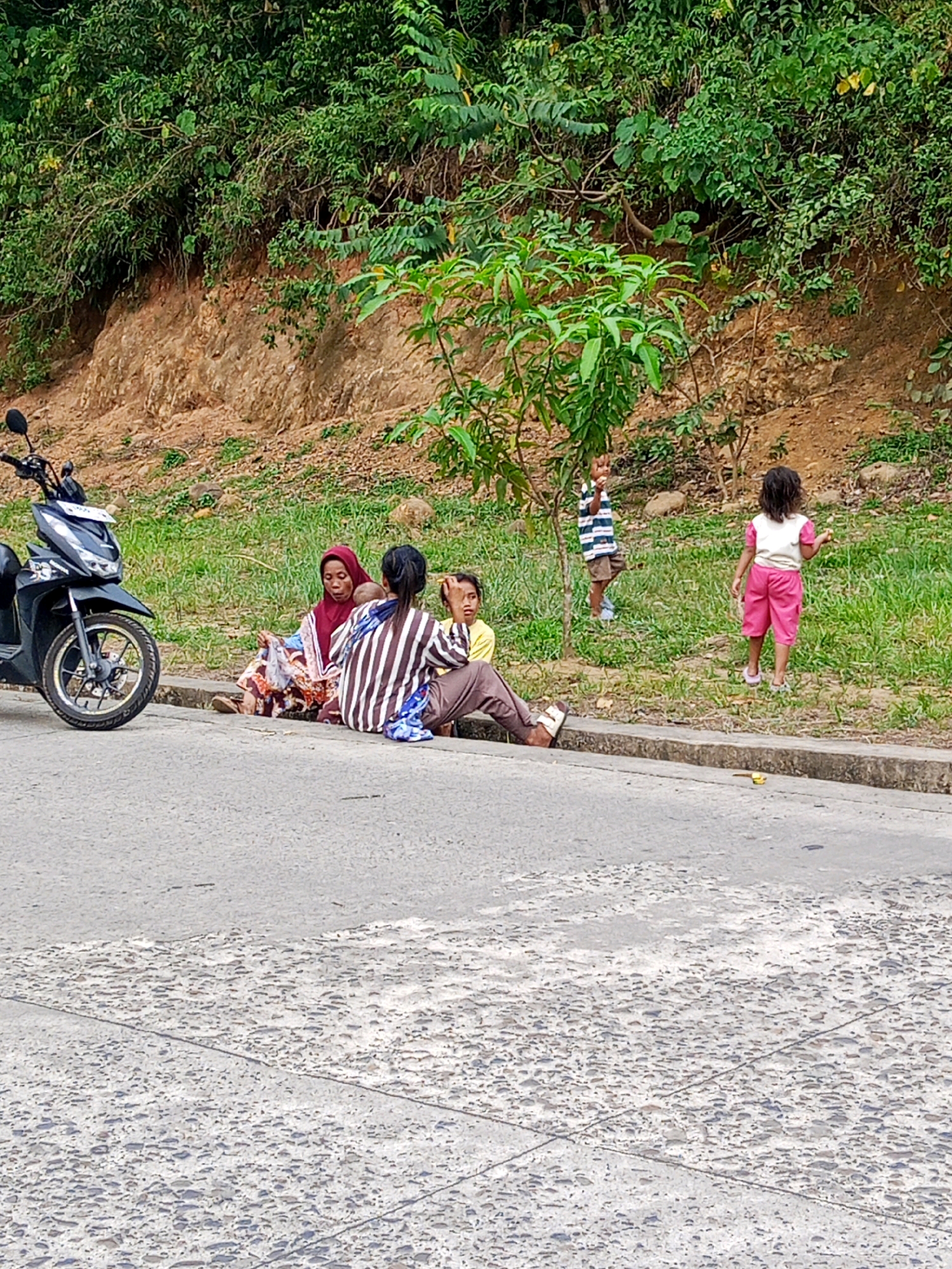 On a paved roadside in Lubuklinggau, Indonesia, a group of people are gathered. A black scooter is parked on the left side of the image. In the center, three individuals are sitting on the curb: an adult female wearing a red hijab and a patterned dress, holding an infant; another adult, possibly female, wearing a striped long-sleeved shirt and brown pants; and a child with dark hair and wearing a yellow shirt. To the right, two young children are standing on the grassy verge. One child, wearing a striped shirt and shorts, is standing and facing away from the camera with their arm raised. Another child, wearing a light-colored shirt and pink shorts, is also facing away from the camera and appears to be walking. The background consists of a steep, vegetated embankment.