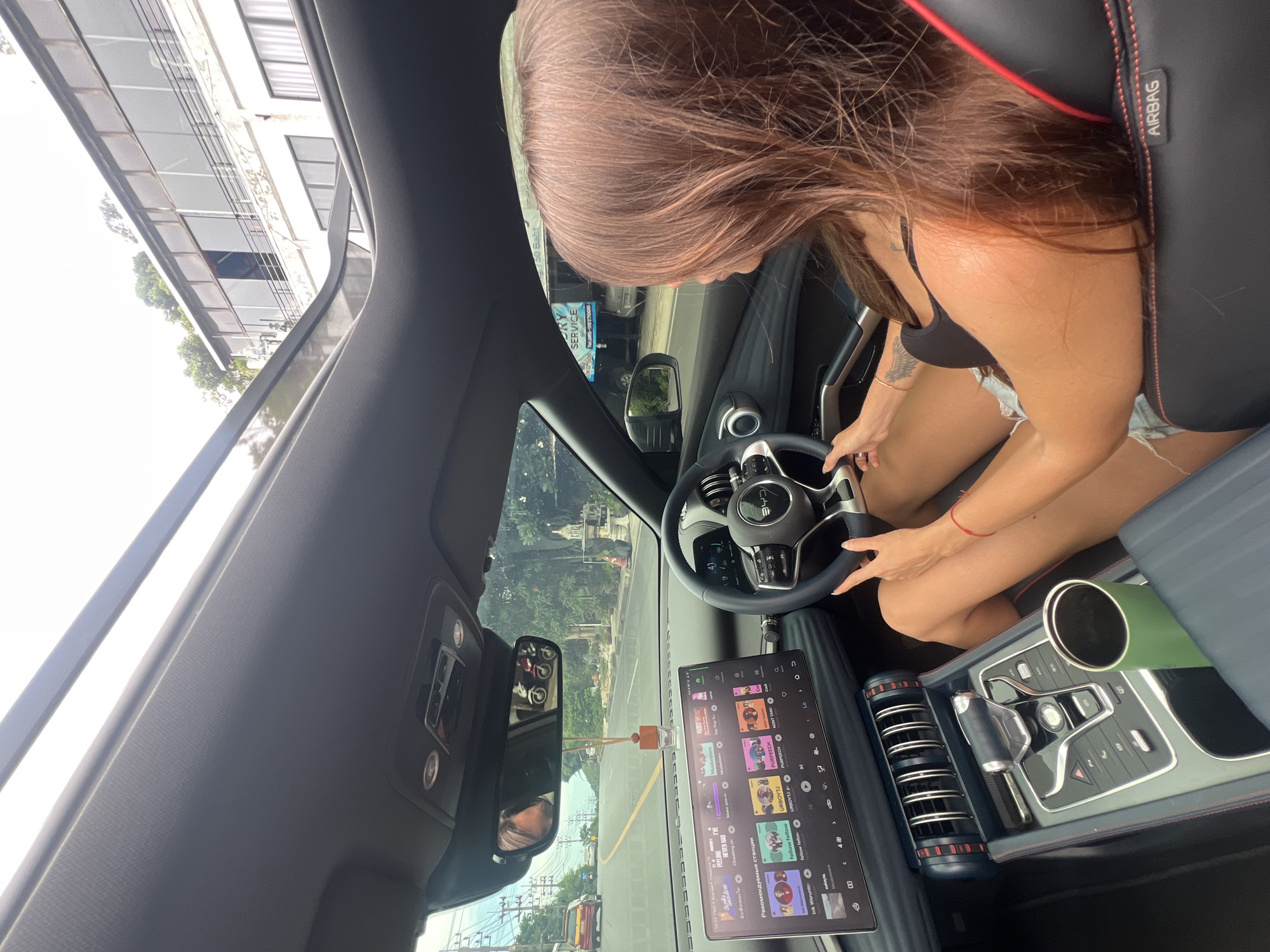 A woman with long, brown hair is driving a car with a sunroof open, looking out at a street in Baan Chaweng Noi, Thailand. She is wearing a black tank top and denim shorts, with a red string bracelet on her right wrist and a tattoo on her left arm. The car's dashboard displays a large touchscreen with music playing. The road outside is paved, with trees and buildings visible on either side. The sky is cloudy.